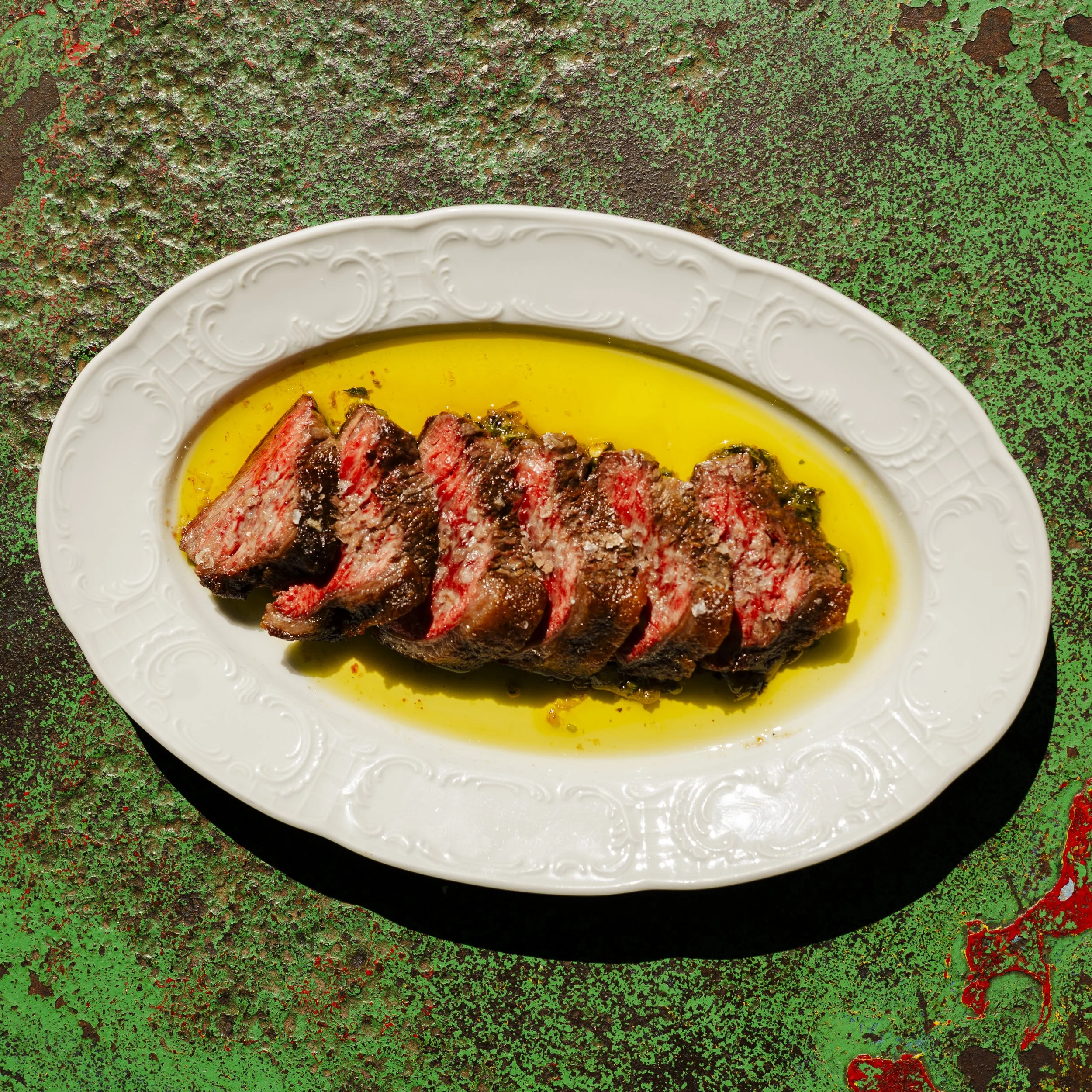 Close-up of a white ornate plate with sliced medium-rare steak with a pink center and a golden oil or sauce. The plate is on a textured green, red, and brown surface.