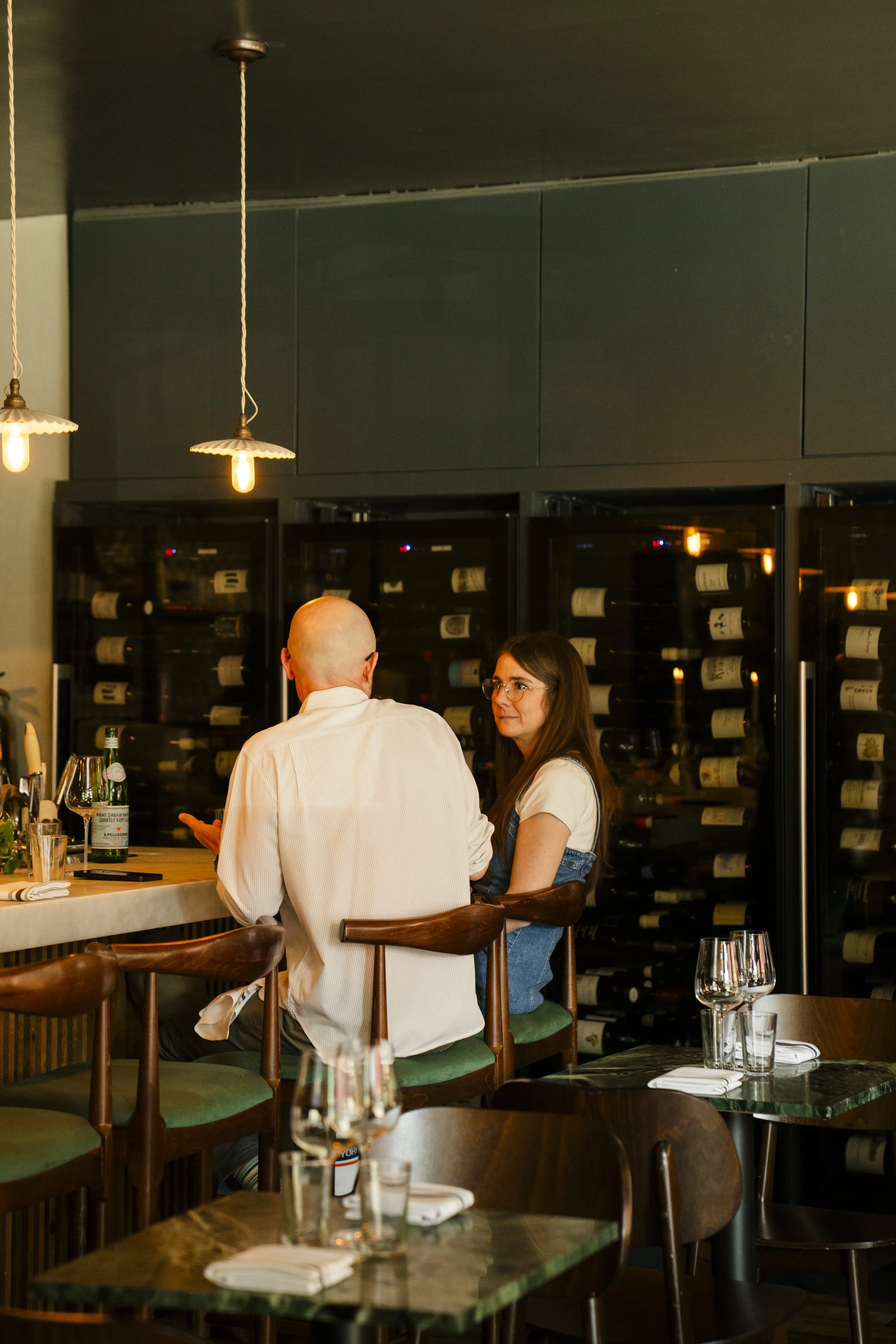 A man and a woman sitting at a restaurant bar, talking with glasses of wine and bottles behind them, illuminated by pendant lighting.