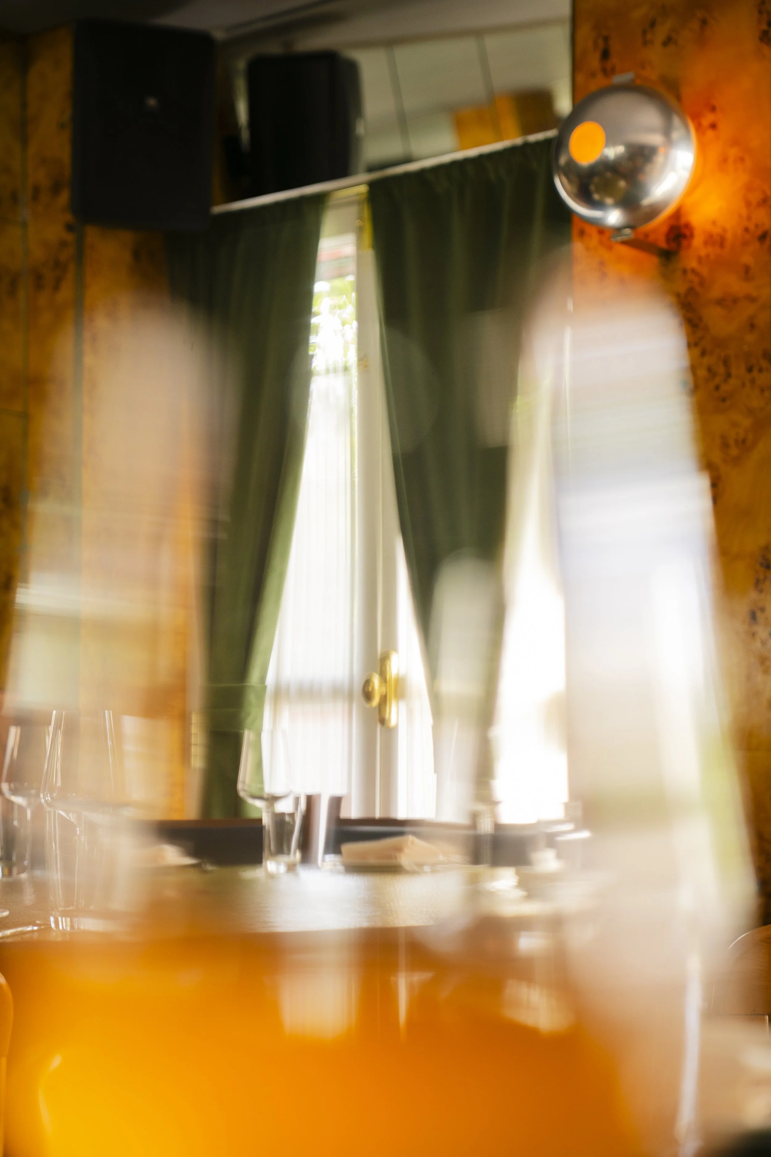 Blurred image of a restaurant or dining area with dark green curtains, a window, and a reflective wall or mirror. There are wine glasses on the table, and a metallic dish or bowl is mounted on a wooden wall. The photo appears to be taken from a low a
