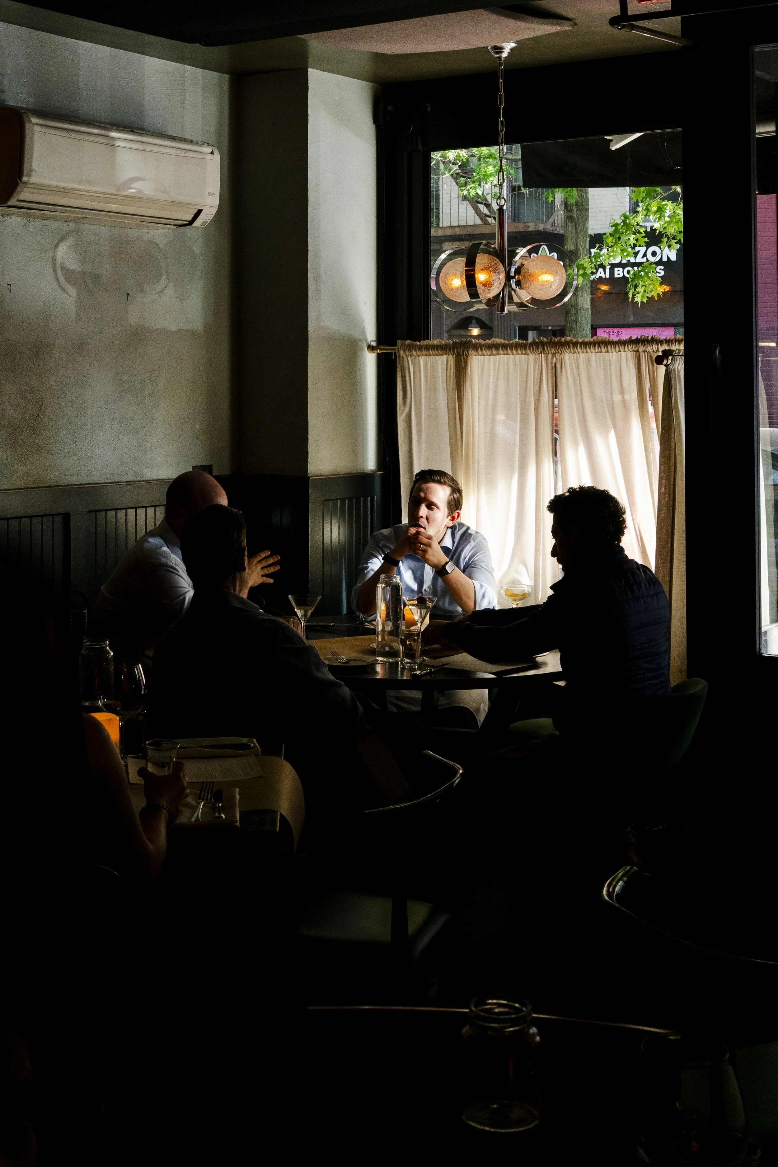 A group of five people sitting around a table in a dimly lit restaurant, with sunlight streaming in through the window behind them. A hanging light fixture is visible above them.