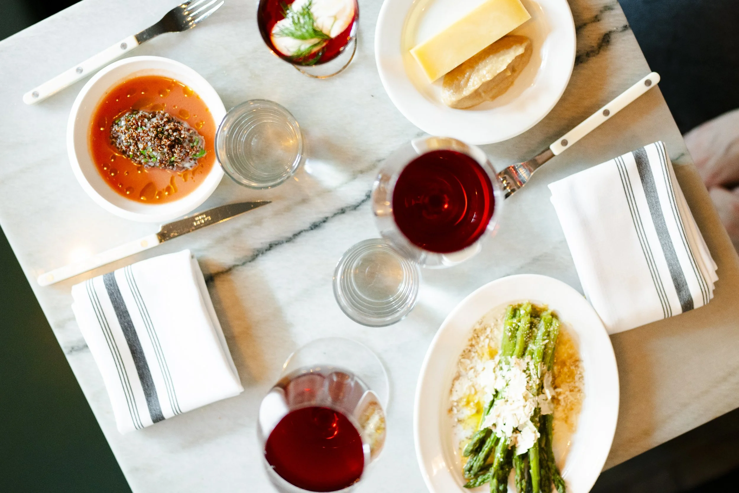Restaurant table setting with plates of pasta, a glass of red wine, a glass of water, and a bowl of tomato-based appetizer. White napkins with black stripes are placed on the table.