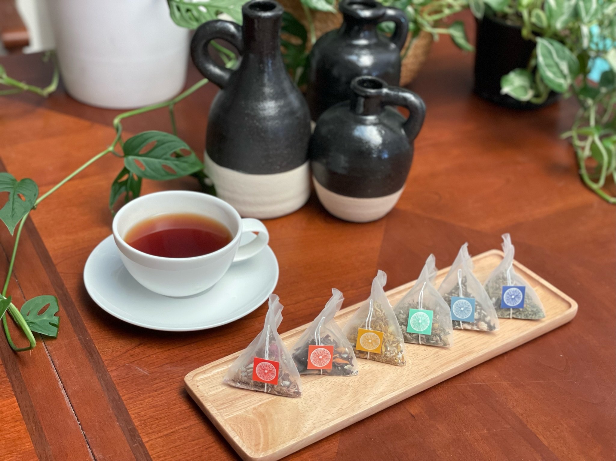 Set of three black and white ceramic vases with handles on a wooden table, surrounded by green plants, a white cup of tea, and a wooden tray with colorful tea sachets.