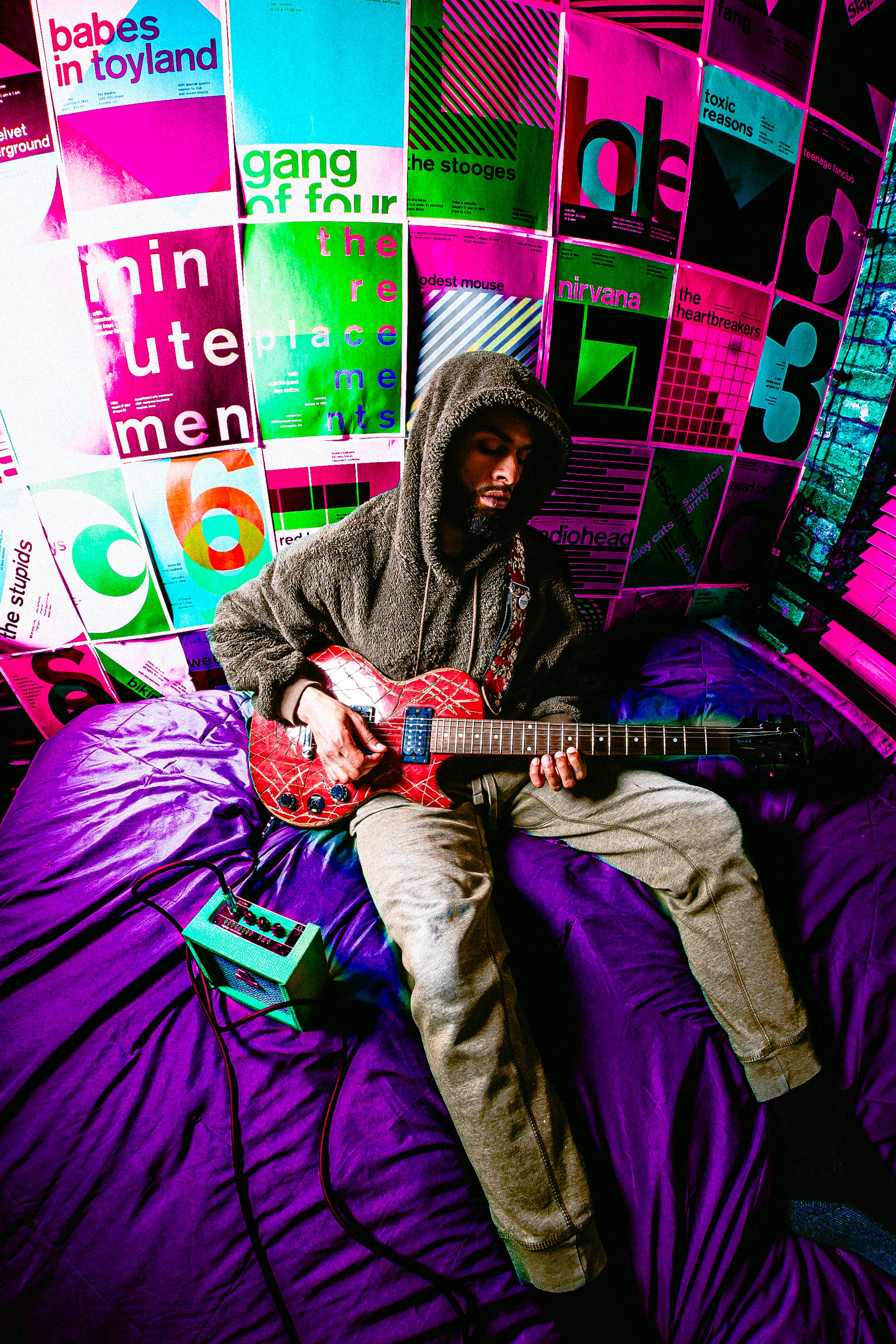 A man sitting on a purple bed in a room with colorful posters on the wall, playing an electric guitar. The posters feature band names and designs, with neon colors and bold text.