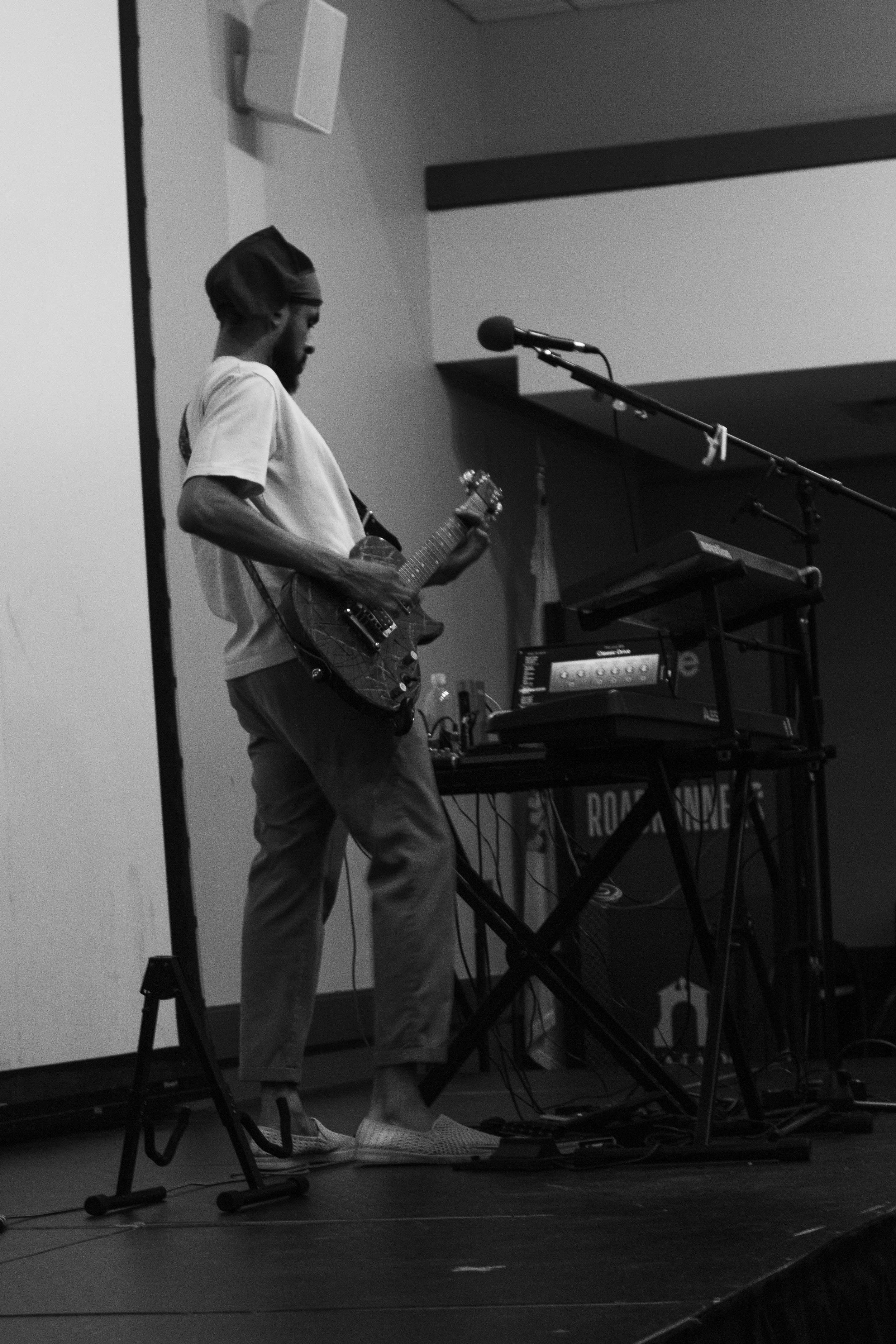 A man wearing a cap, white T-shirt, and sneakers is playing an electric guitar on a stage with music equipment, including a keyboard and a microphone.