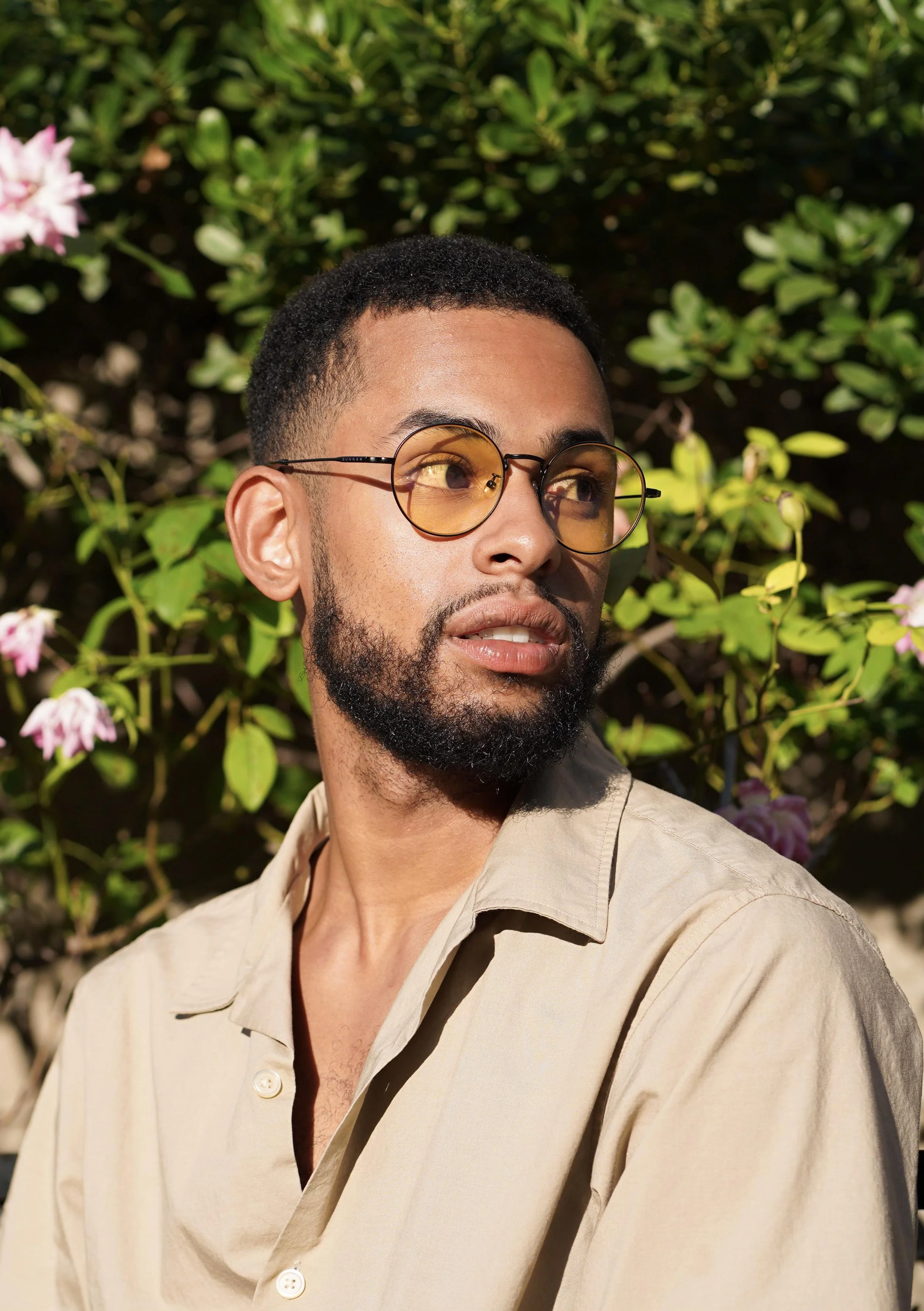 A man with short curly black hair and a beard is wearing round yellow-tinted sunglasses and a beige shirt, looking to his right. He is outdoors with greenery and pink flowers in the background.