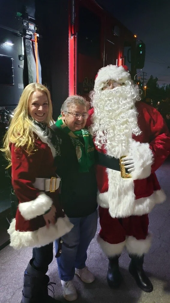Three people, two women and a man, posing with Santa Claus at night. The women are dressed in Christmas-themed outfits, and Santa is in his traditional red suit with a white beard.