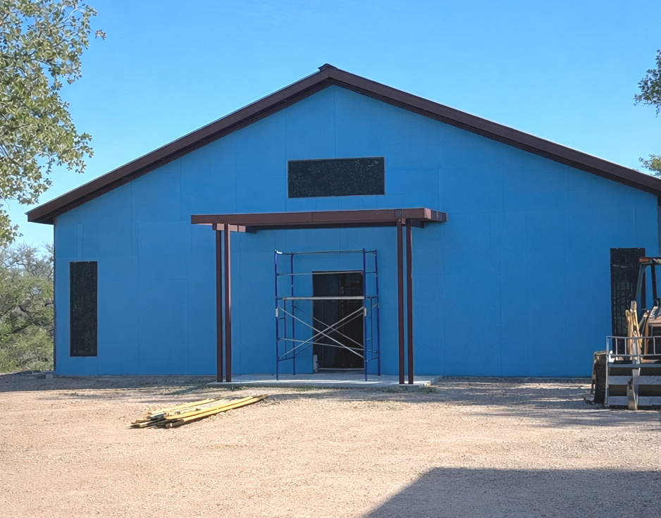 A building under construction painted blue with scaffolding in front, a small entrance with a red frame, and construction equipment on the right side.