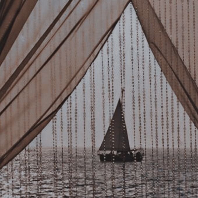 A sailboat on the water seen through a decorative beaded curtain with a triangular opening.