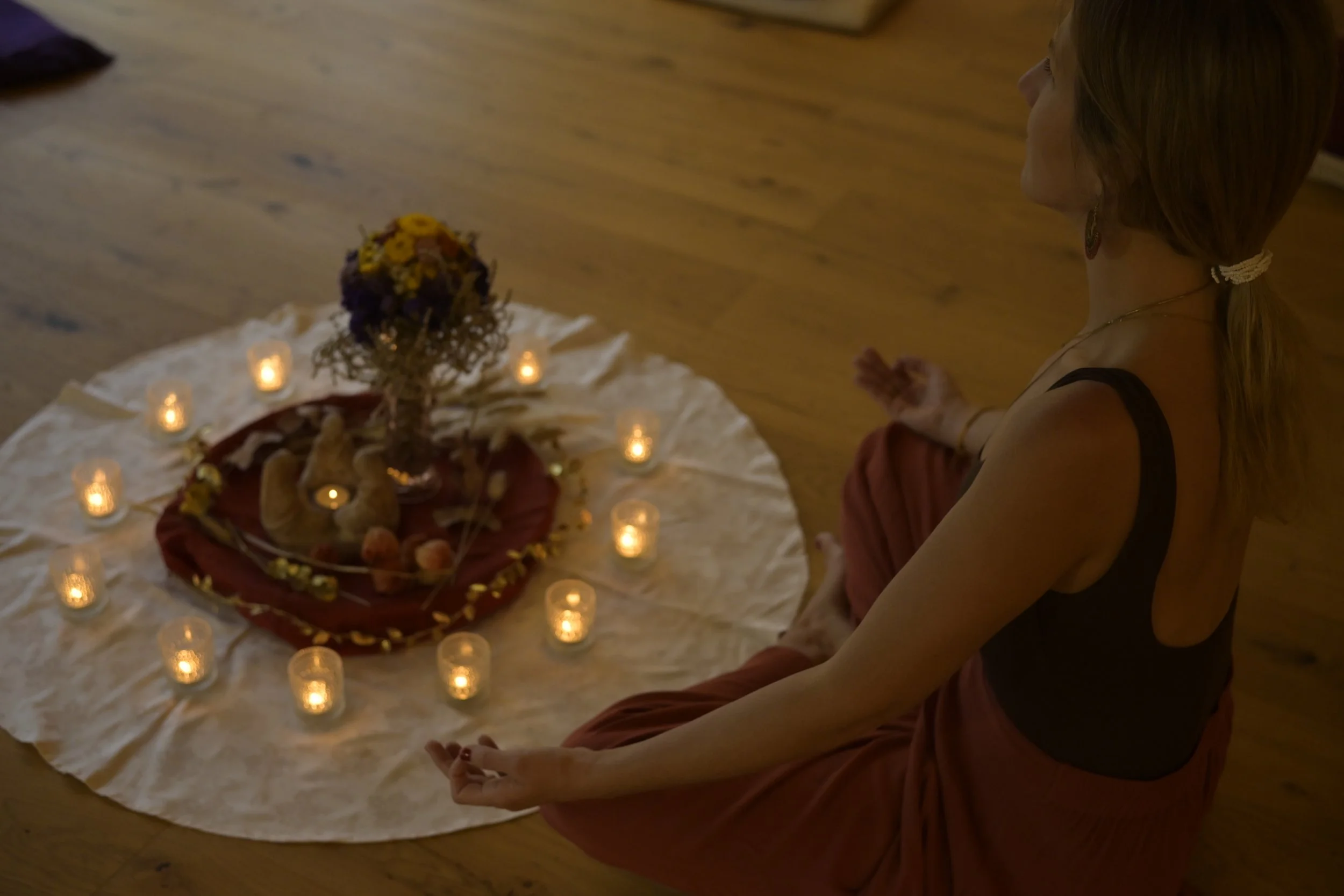 A woman with long hair wearing a dark dress and jewelry is sitting cross-legged on the floor with her hands in a prayer position in front of a decorated altar with a floral arrangement and candles.