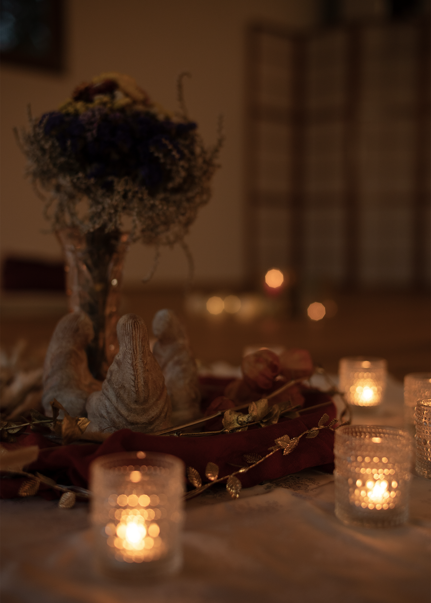 A dimly lit decorative display with dried flowers, candles in glass holders, and a garnet-colored fabric with gold accents, creating a warm and cozy ambiance.
