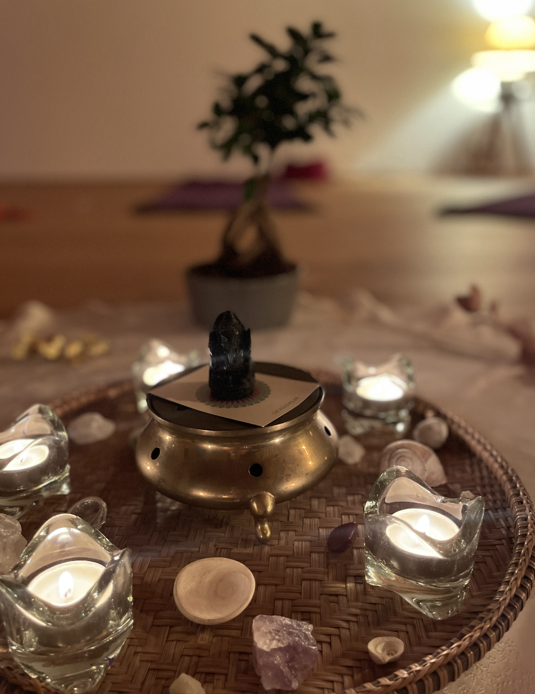 A small oil burner with a lit candle on top, surrounded by glass candle holders and seashells, on a round woven tray; a potted plant and lamp in the background.
