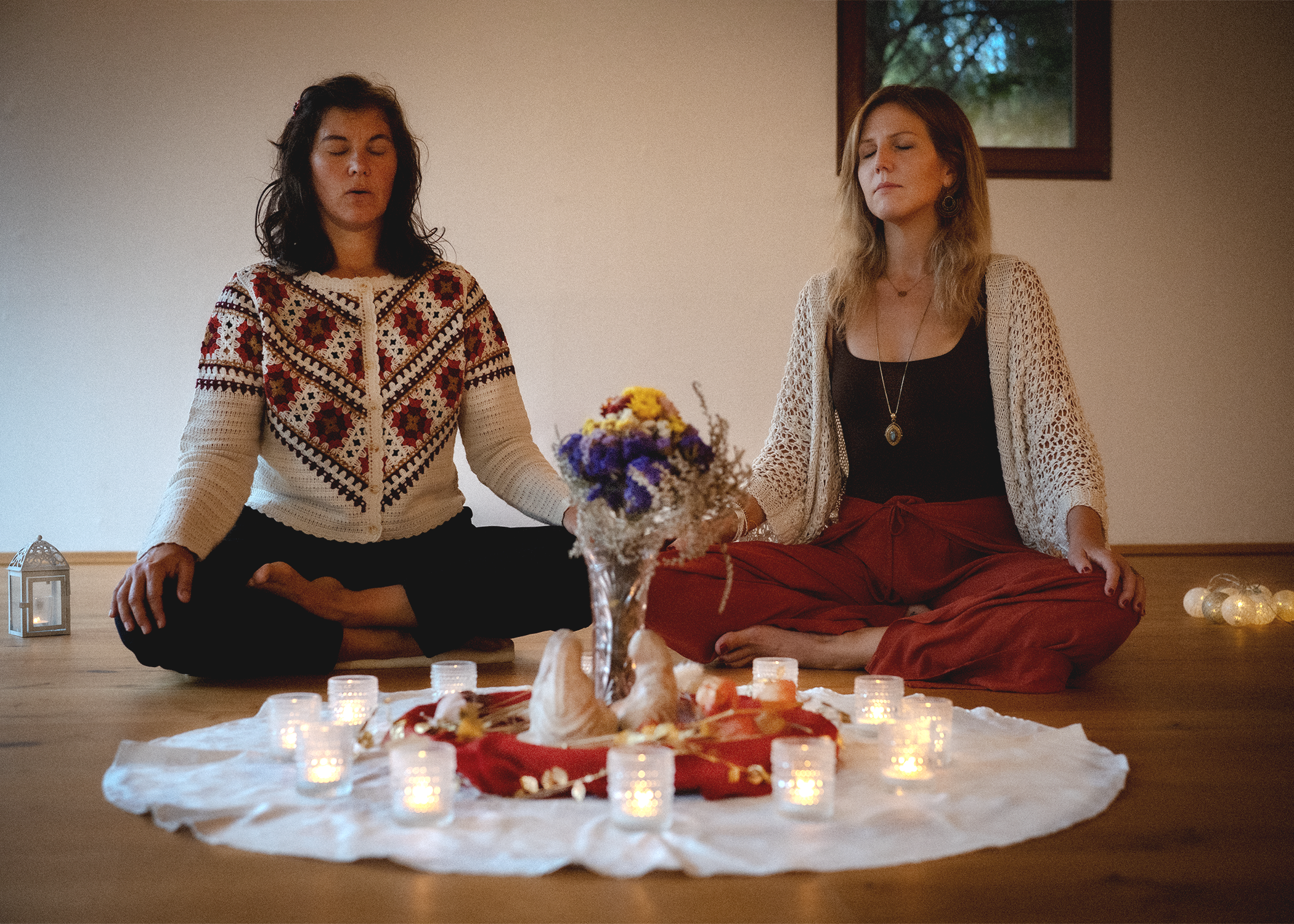 Two women in meditation surrounded by candles, flowers and ritual objects. It is a women circle they are holding. 
