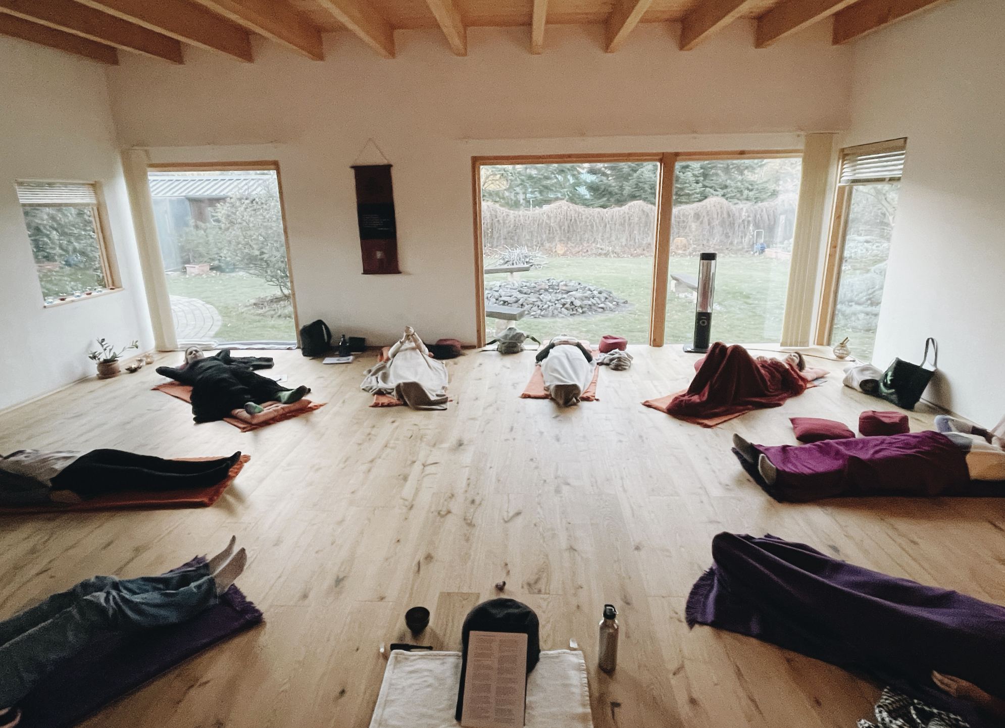 A group of people practicing yoga or meditation in a spacious room with large windows and wooden ceiling beams. They are lying on mats on the wooden floor in a relaxed position, facing the windows overlooking a garden.