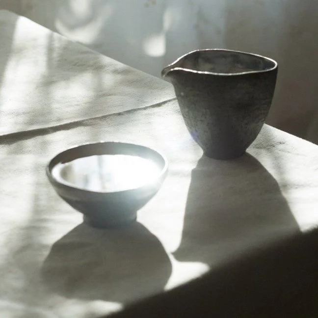 A small metal bowl and a larger ceramic pitcher on a wooden surface, with shadows cast by sunlight.