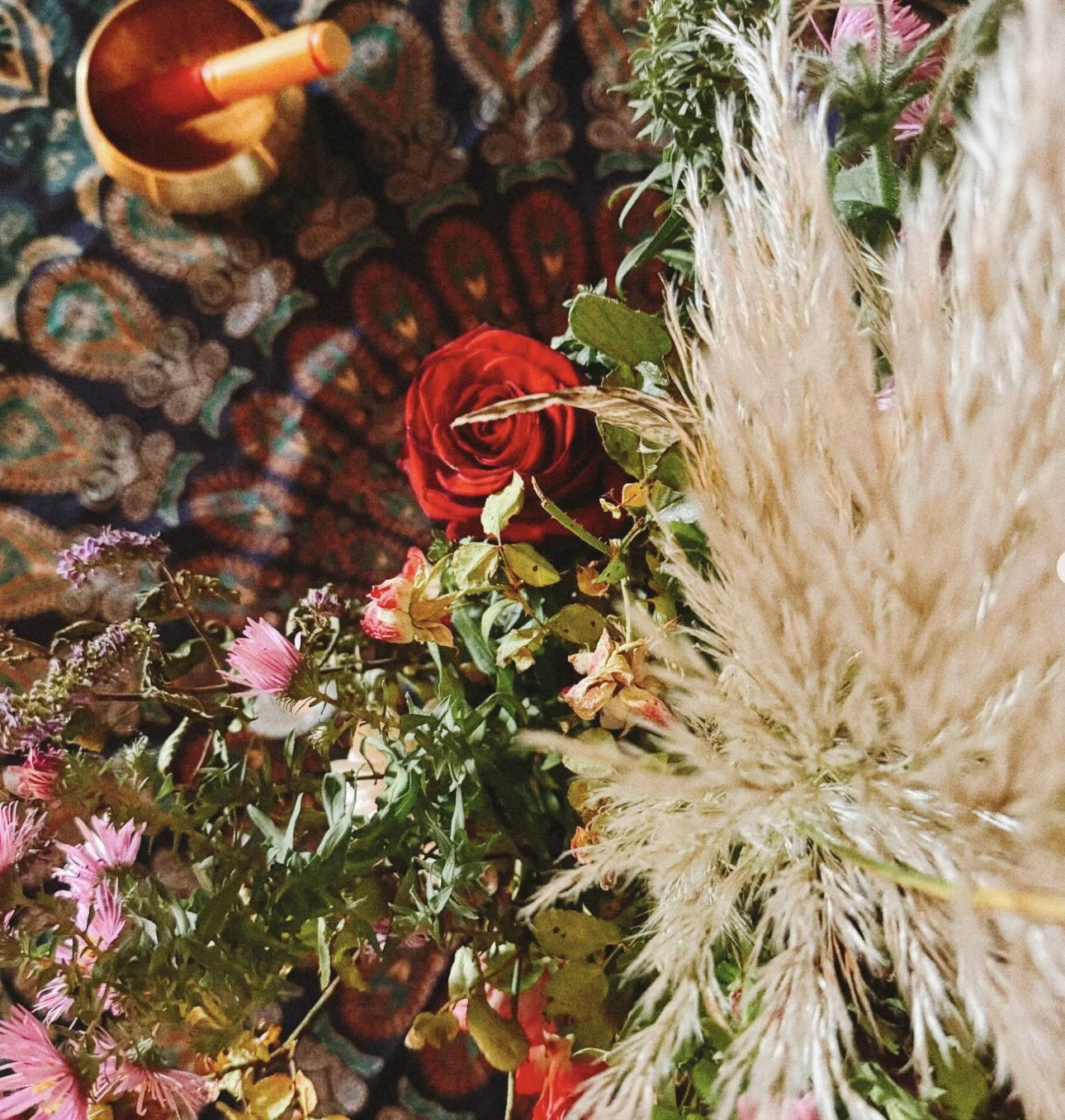 Arrangement of flowers including a red rose, pink asters, and beige pampas grass on a patterned fabric background.