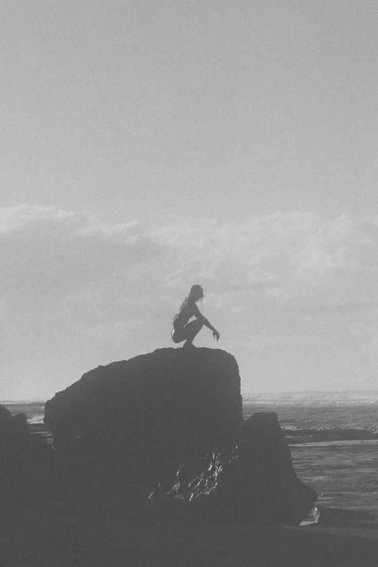 A person crouching on top of a large rock by the ocean during daytime, with cloudy sky in the background.
