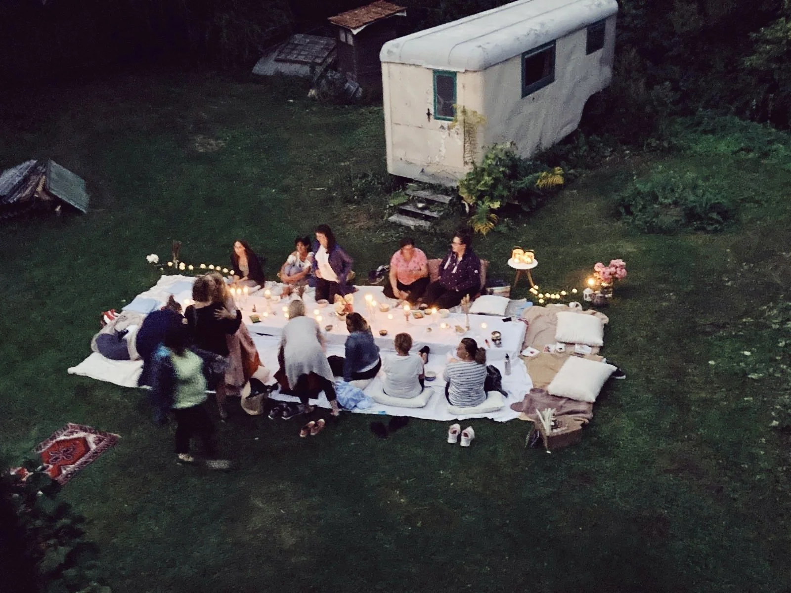 People gathered around a low table outdoors for a picnic or celebration, with candles and decorations, in front of a vintage trailer, at dusk or nighttime.