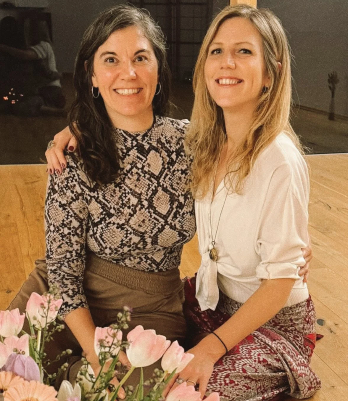 Two women smiling and sitting close together at a table with pink flowers. One woman has dark curly hair, wearing a patterned top, and the other has blonde straight hair, wearing a white blouse. They are indoors with a wooden floor and a window in th