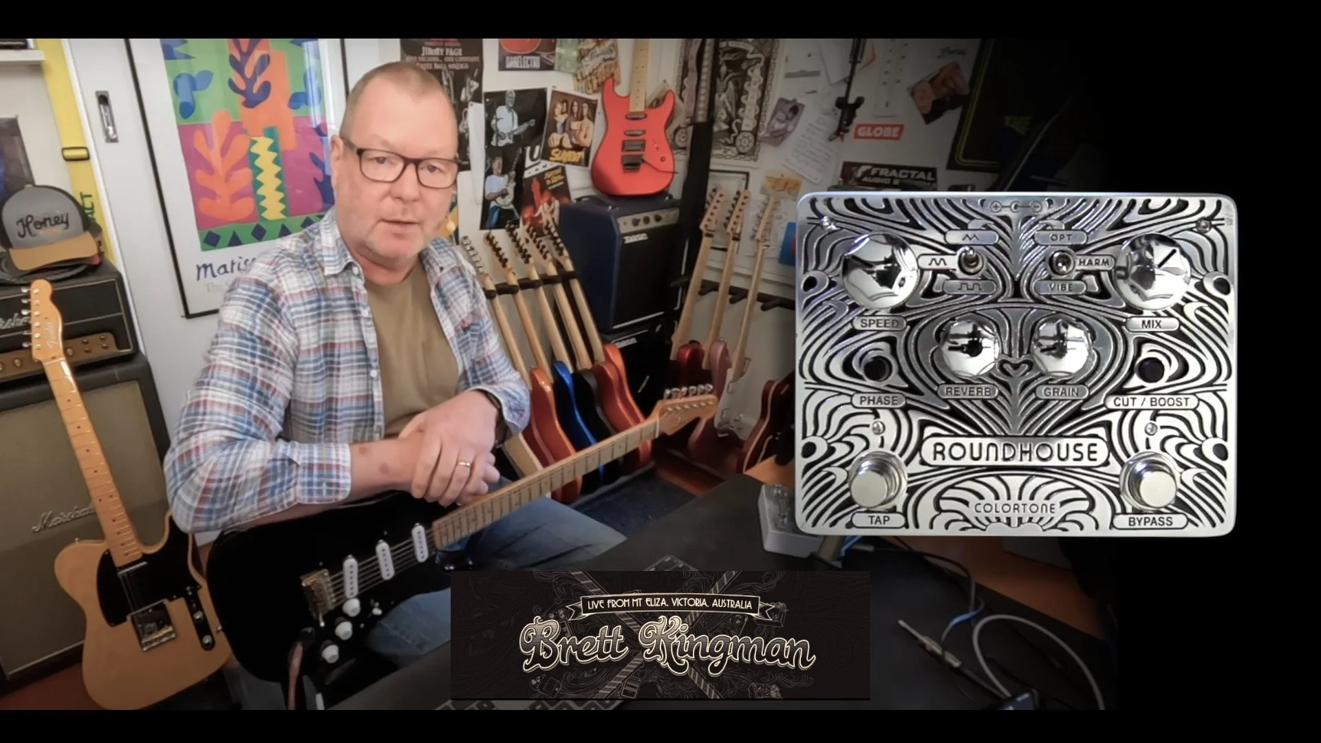 A man wearing glasses and a checkered shirt sitting with an electric guitar in a music studio filled with guitars on racks and amplifiers. There are posters on the walls and a close-up of a guitar effects pedal labeled 'Roundhouse'.