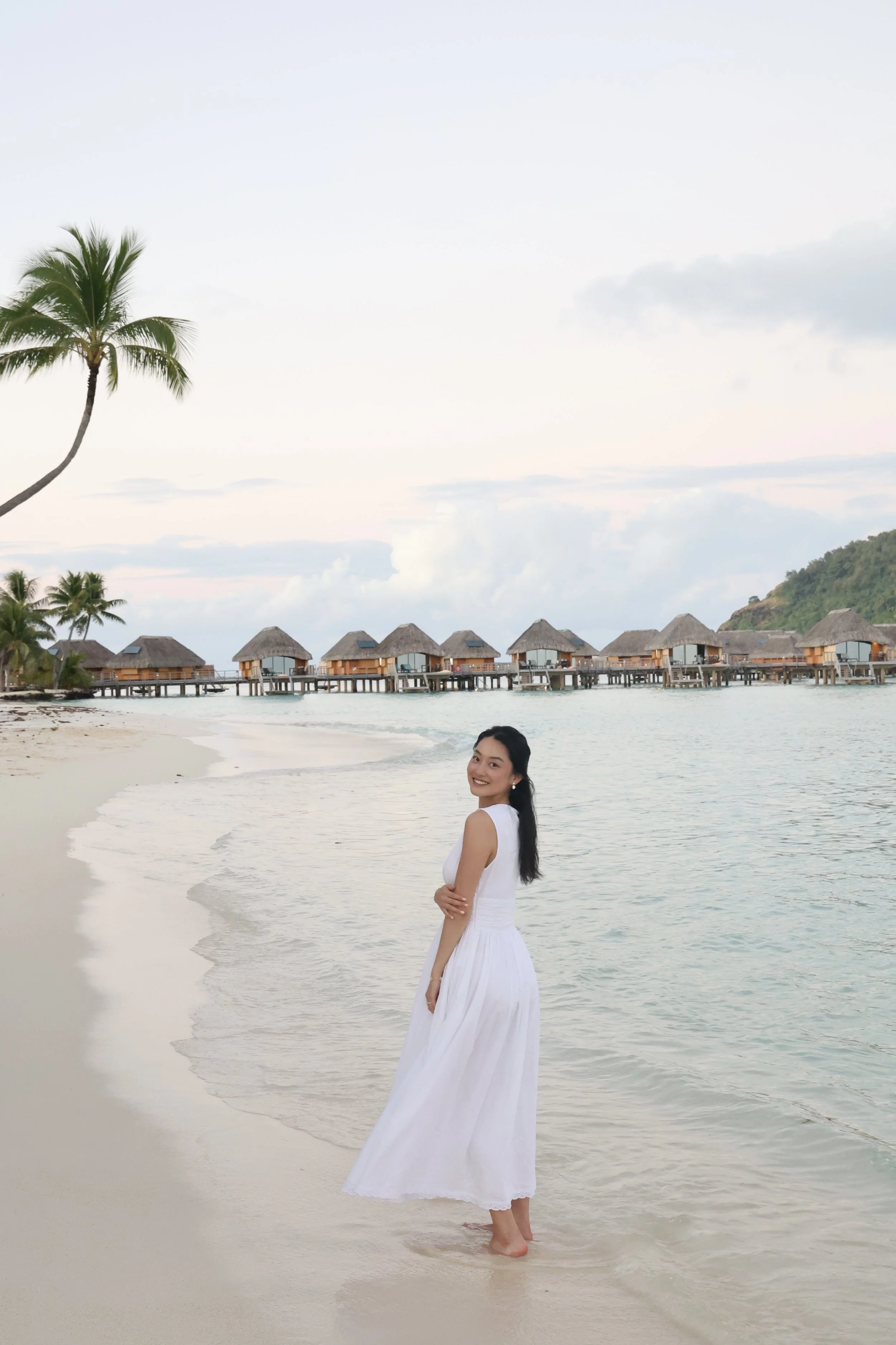 A woman in a white dress standing barefoot on the beach near the water, with overwater bungalows in the background, and a palm tree on the left side of the image.