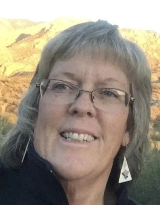 Close-up of a smiling woman with glasses and earrings, outdoors with a mountainous landscape in the background.