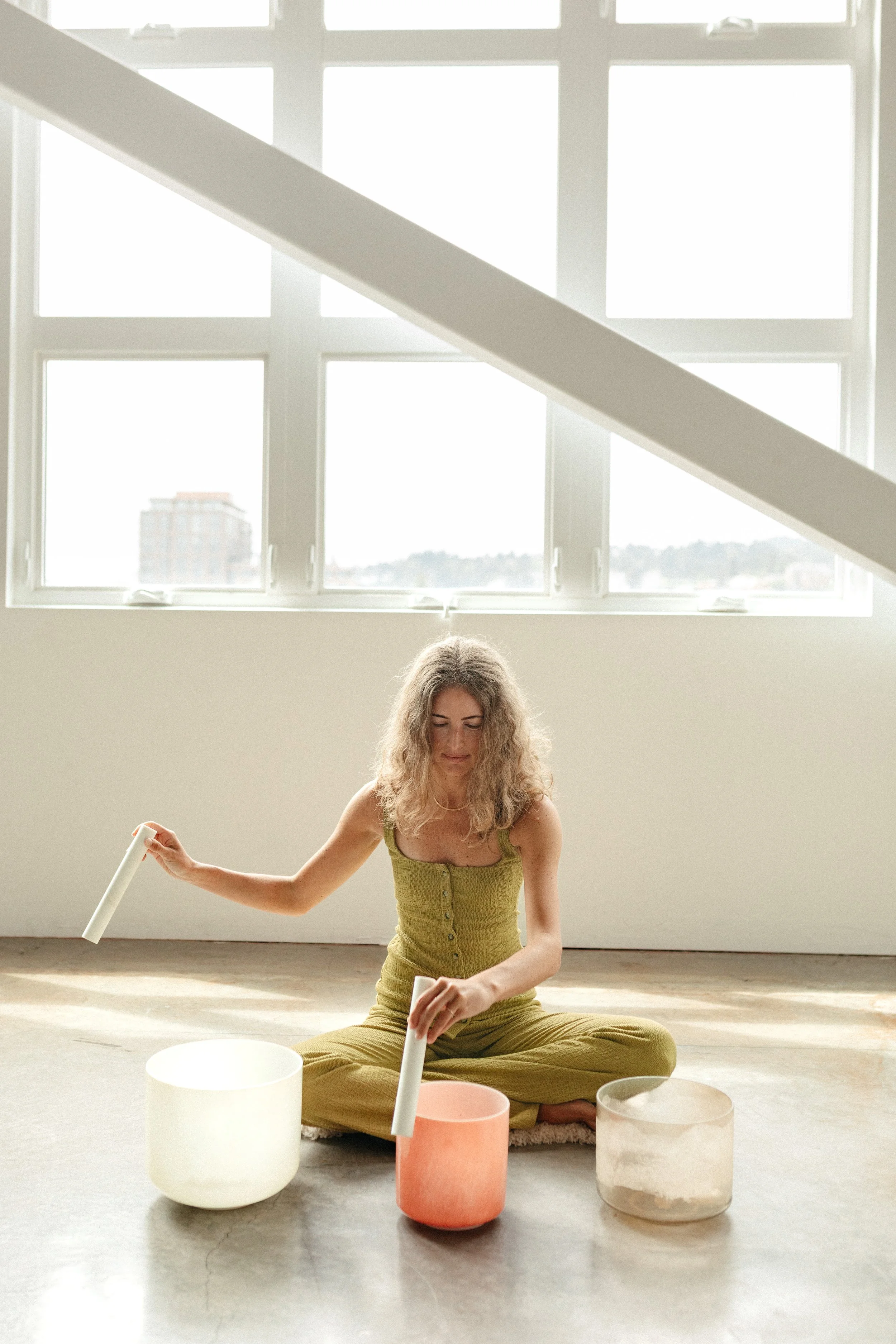 A woman sitting cross-legged on the floor playing crystal singing bowls in a bright room with large windows.