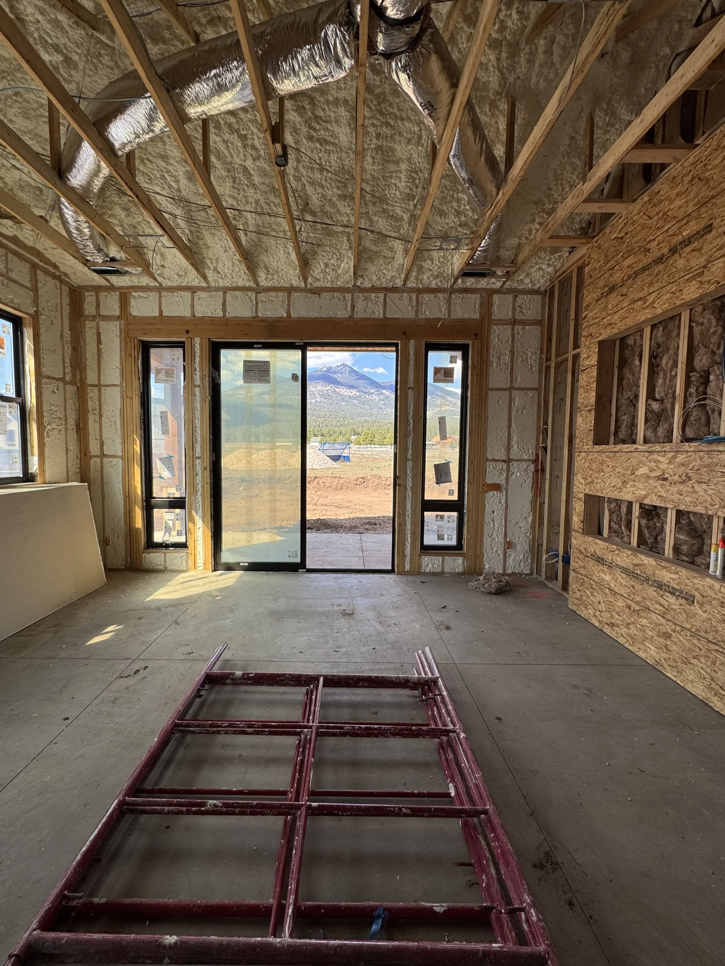 Interior of a house under construction with unfinished walls and ceiling insulation, a sliding glass door leading outside with mountain views, and construction equipment on the floor.