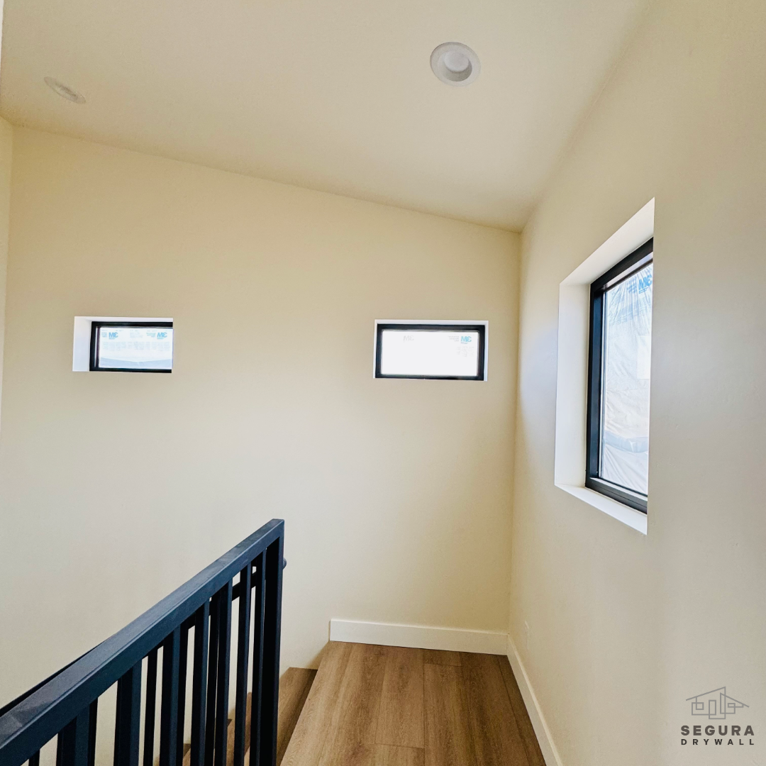 Interior view of a staircase landing with three small rectangular windows, beige walls, wooden flooring, black railing, and a ceiling light