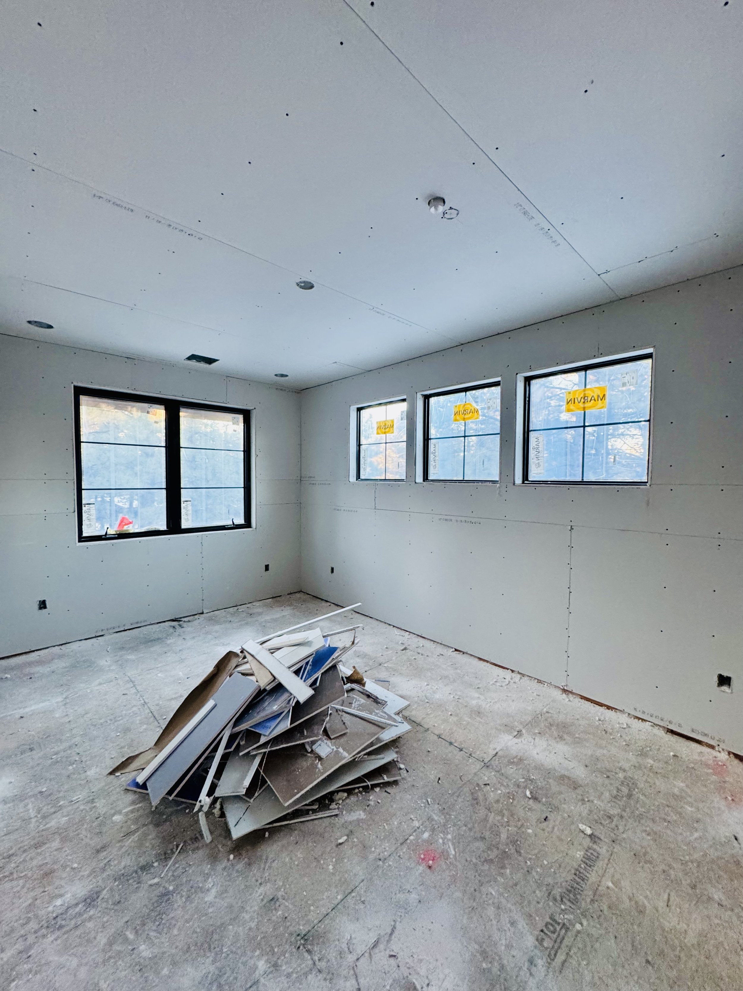 An unfinished room under construction with drywall installed, three windows on one wall, and a pile of drywall scraps on the floor.