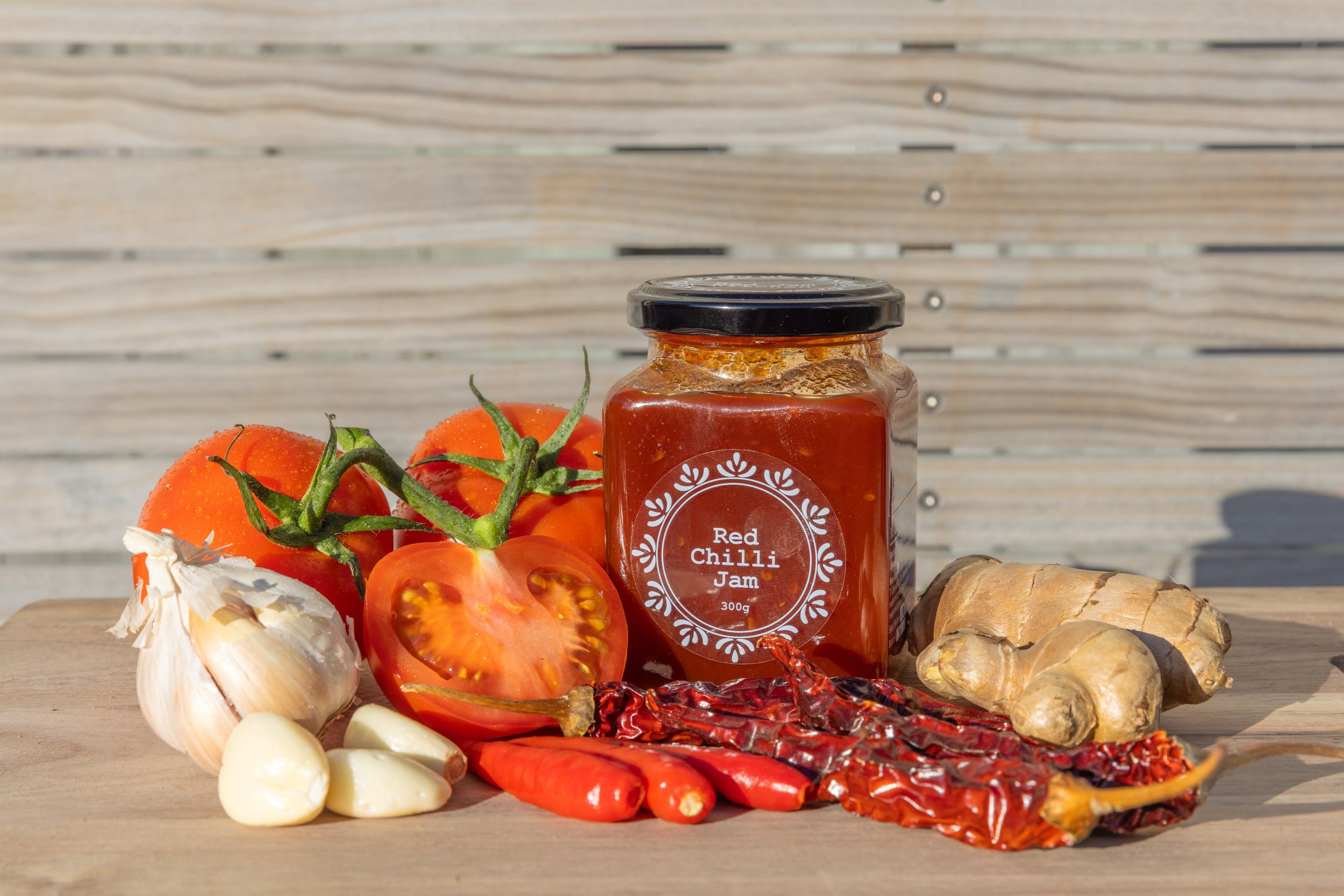 Tomatoes, garlic cloves, ginger, red chili peppers, and a jar of red chili jam on a wooden surface with a wooden background.