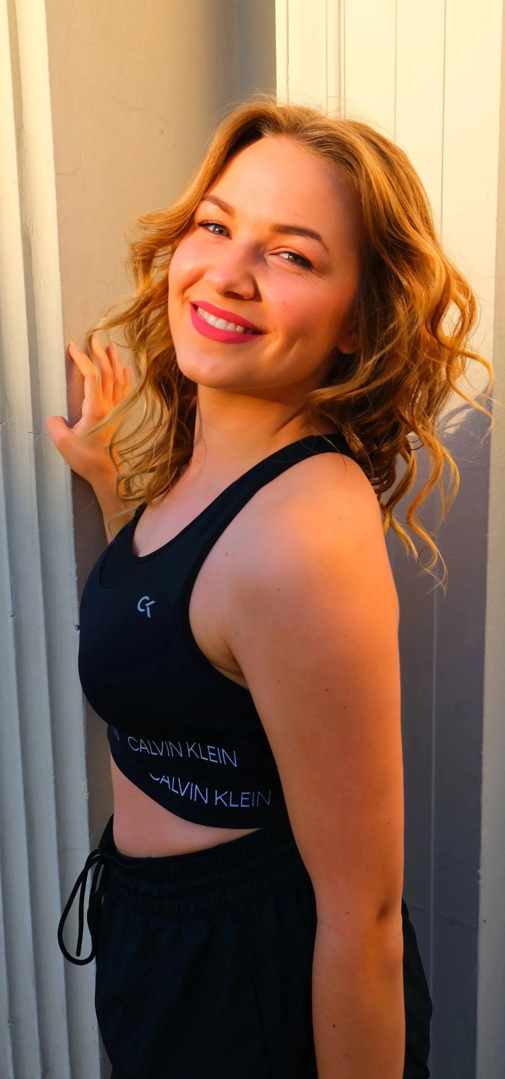 Lächelnde junge Frau mit lockigem, blonden Haar trägt schwarze Sportkleidung, steht neben einer weißen Wand und sieht in die Kamera.