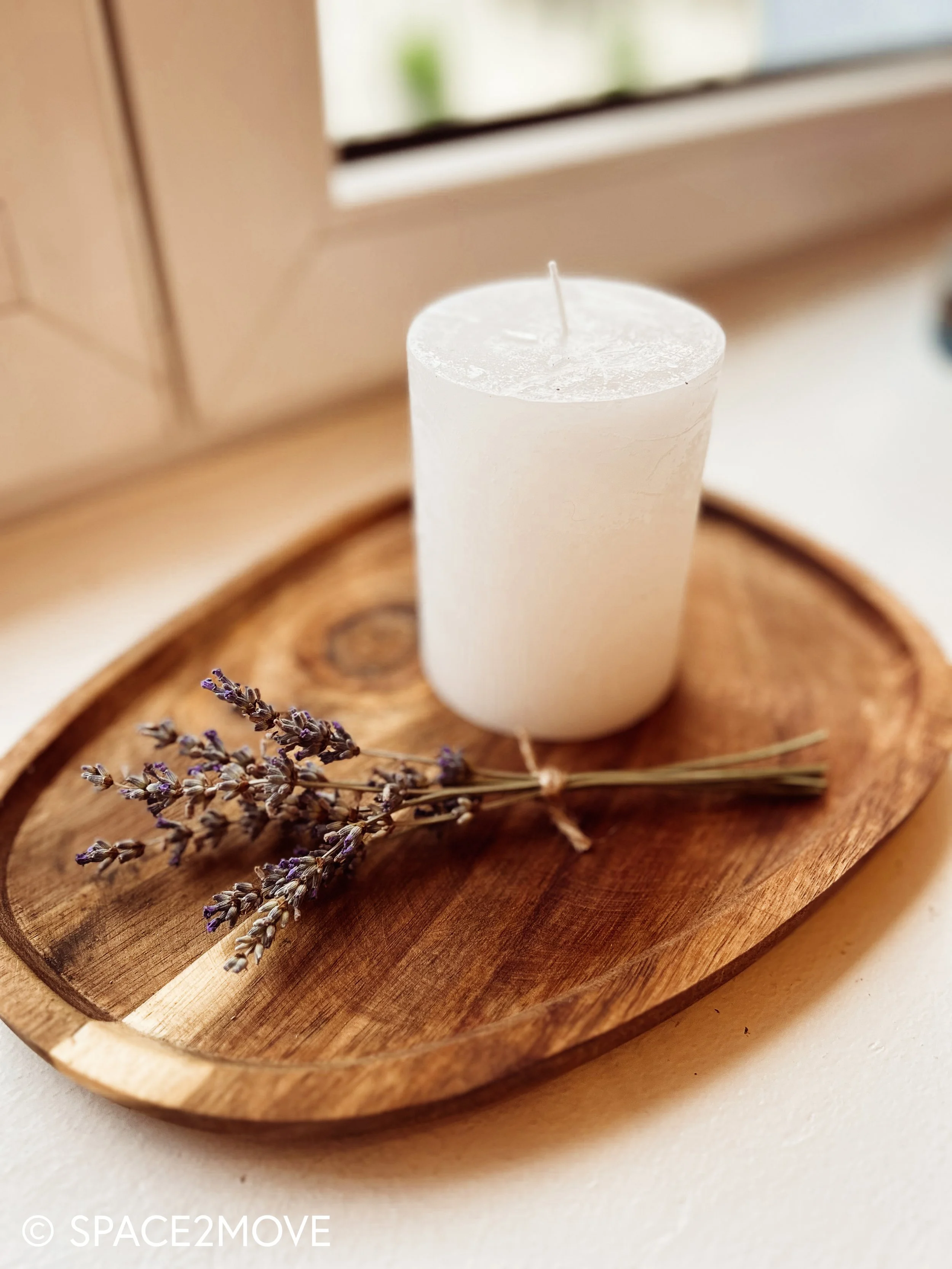 Weiße Kerze auf einem runden Holzteller mit Lavendelsträuchern, vor einem Fenster auf einem weißen Tisch.