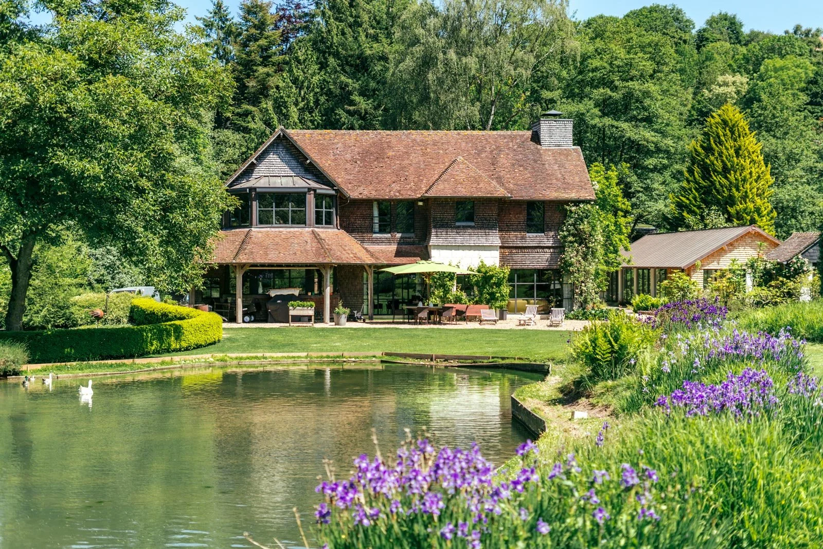 Une maison en bois avec un toit en tuiles rouges, entourée de verdure et de fleurs, avec un étang au premier plan et une forêt en arrière-plan.