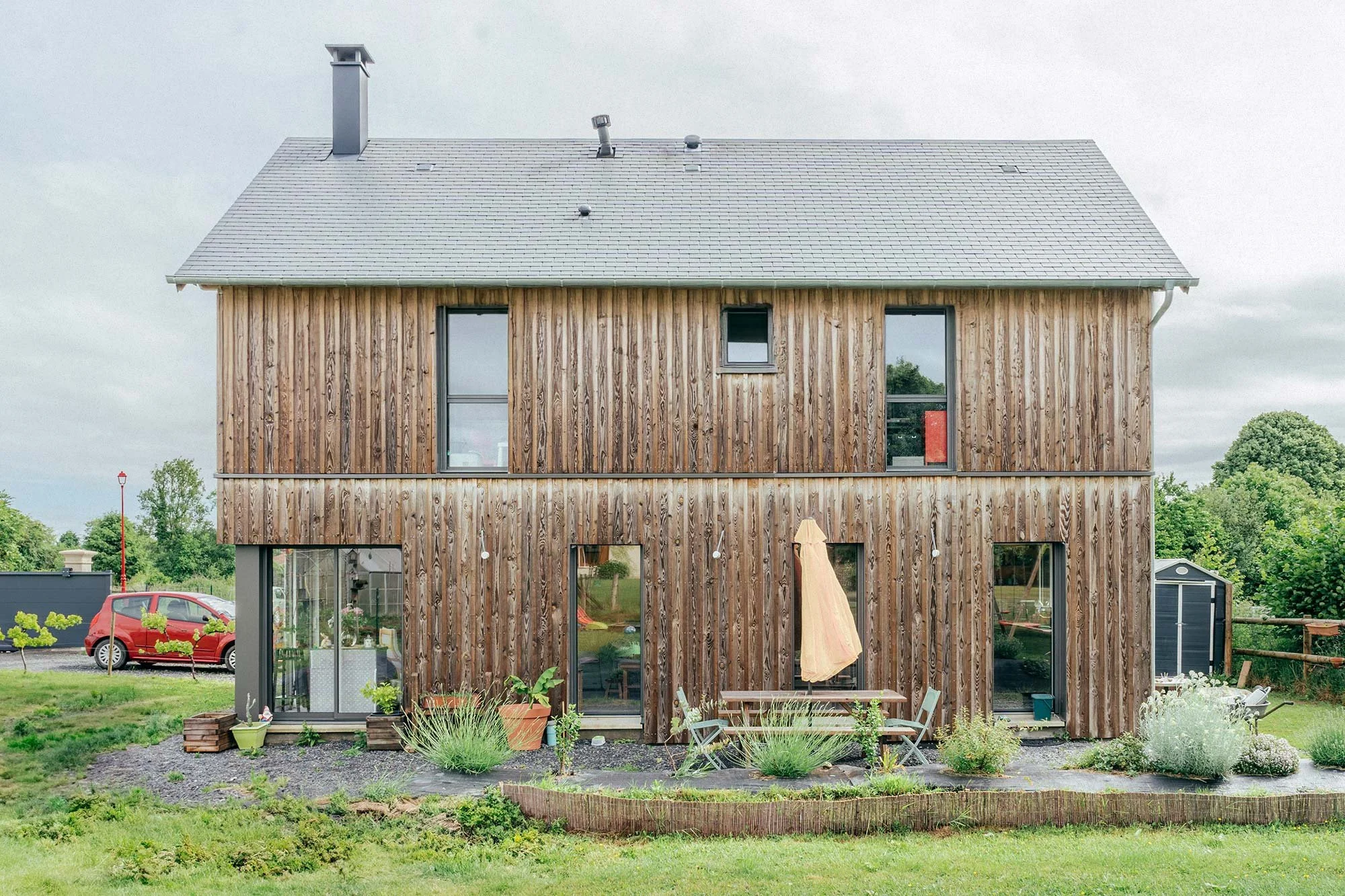 Maison à deux étages avec façade en bois, grandes fenêtres, jardin avec meubles et plantes, arrière-plan avec arbres et ciel nuageux.