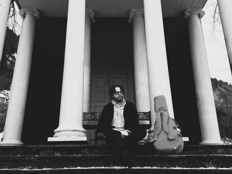 Adran Mork Christoffersen sitting at Mon Plaisir in Bergen with his guitar bag casually leaned against one of the monument's many columns.