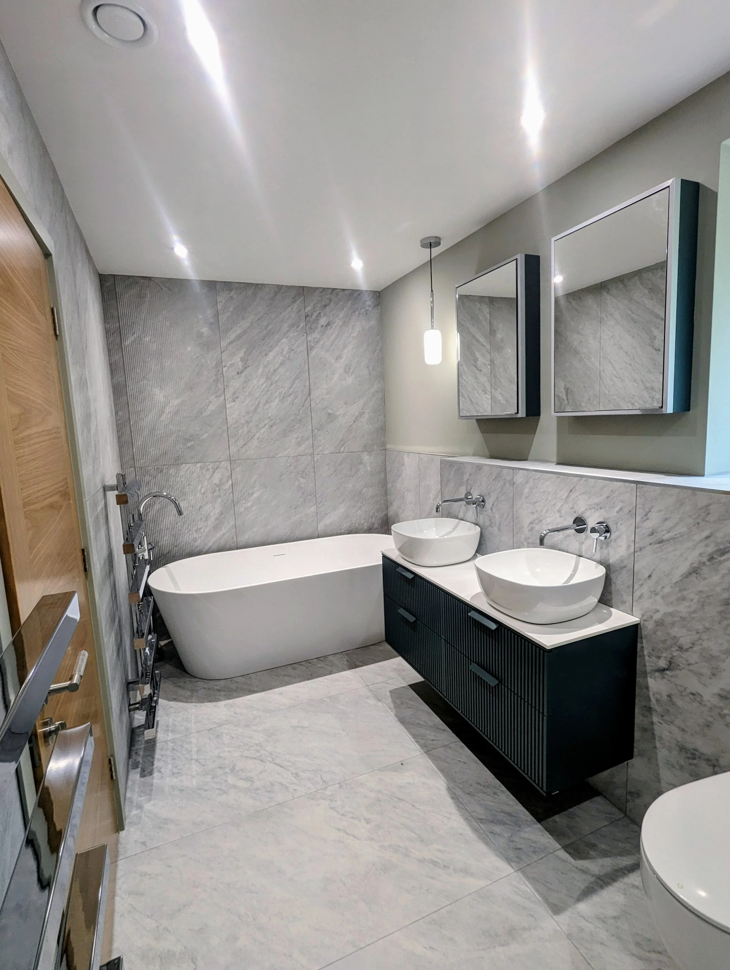 Modern bathroom with a white freestanding bathtub, marble tiles, dual vessel sinks on a black vanity, wall-mounted mirrors, hanging pendant light, and a toilet.