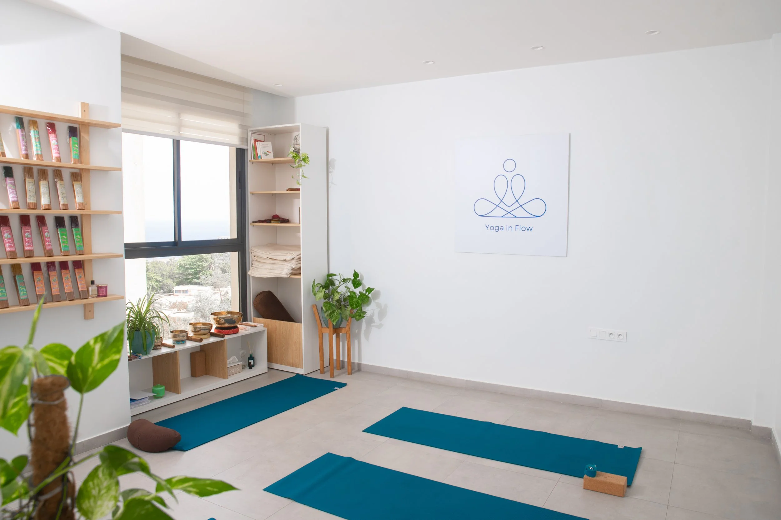 Salle de yoga lumineuse avec deux tapis bleus, plantes vertes, étagère avec des livres et des objets, fenêtre laissant entrer la lumière naturelle, tableau avec le logo 'Yoga in Flow'.