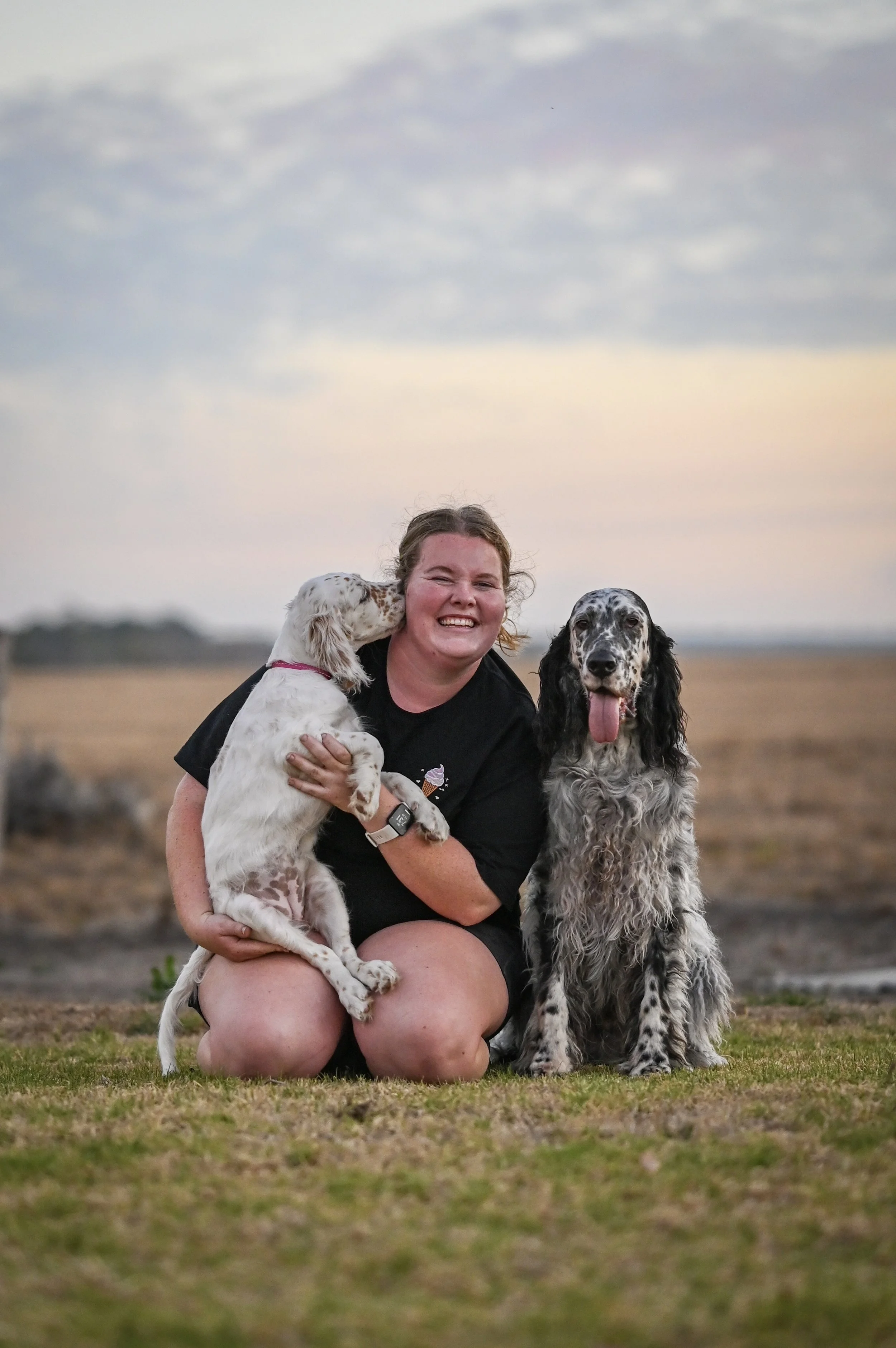 A woman smiling and kneeling on the grass outdoors, holding a white and brown spotted puppy, with a large gray and black speckled dog sitting next to her during sunset.
