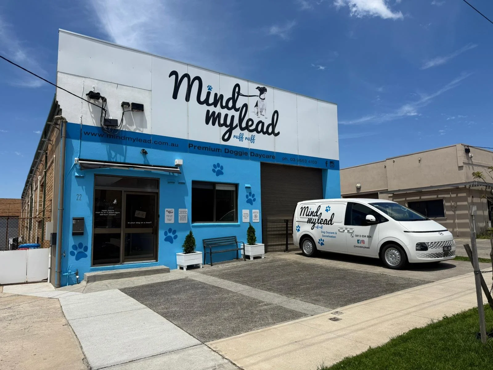 Exterior view of a building with blue walls labeled 'Mind my lead' dog daycare, featuring a large sign with a black and white dog illustration, paw prints, and contact information, with a white van parked in front displaying the business name and paw prints.