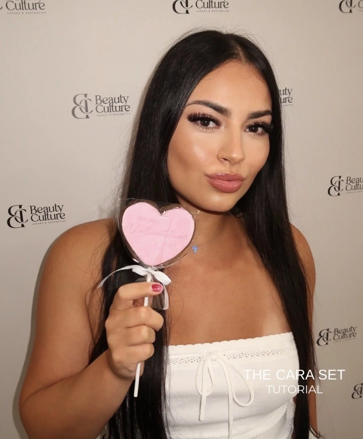 A woman with long dark hair and makeup holding a pink heart-shaped sign. She is wearing a white top and standing in front of a backdrop with 'Beauty Culture' logos. Text on the image reads 'The Cara Set Tutorial.'