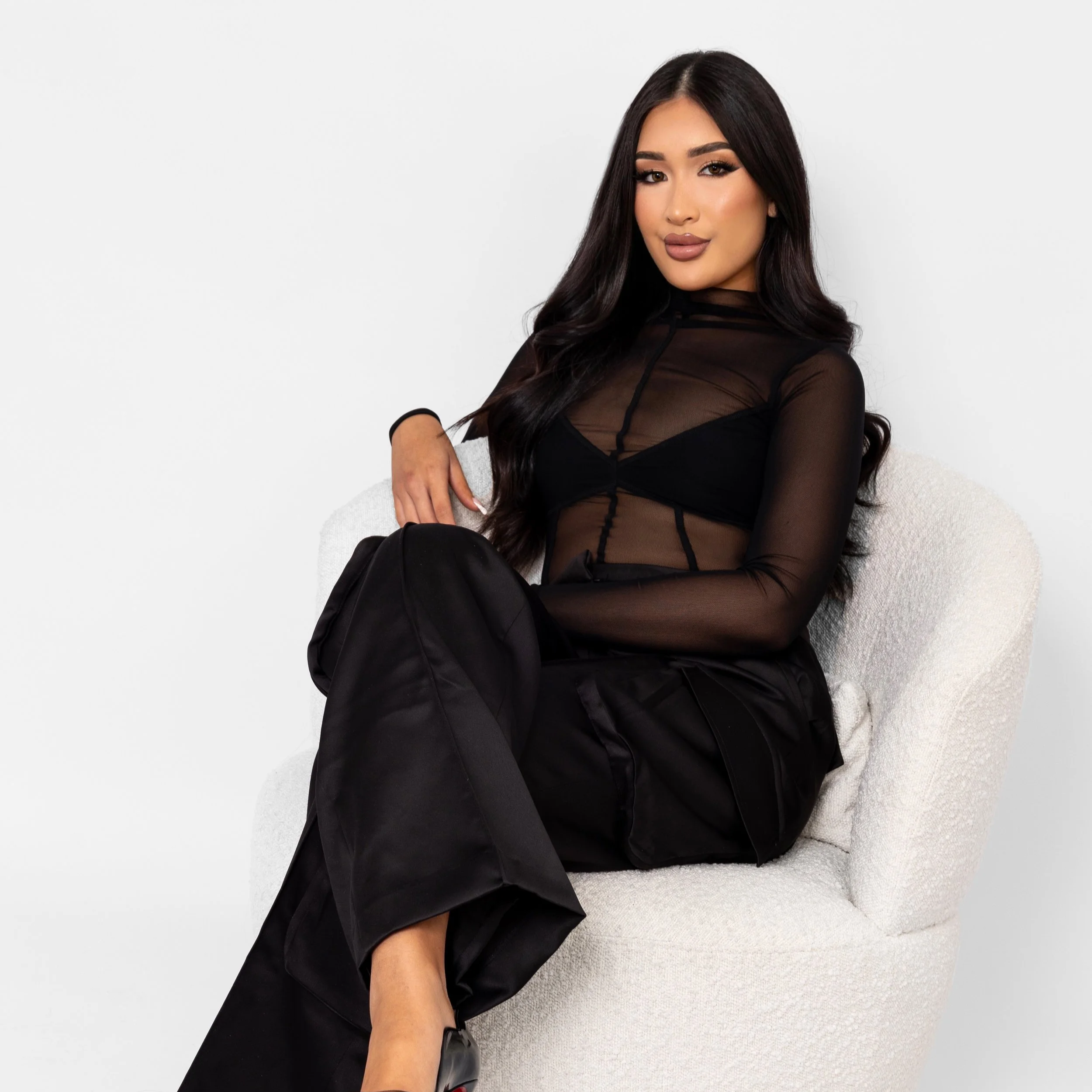 A woman with long dark hair wearing a black sheer top and black pants, sitting on a white textured chair against a plain white background.