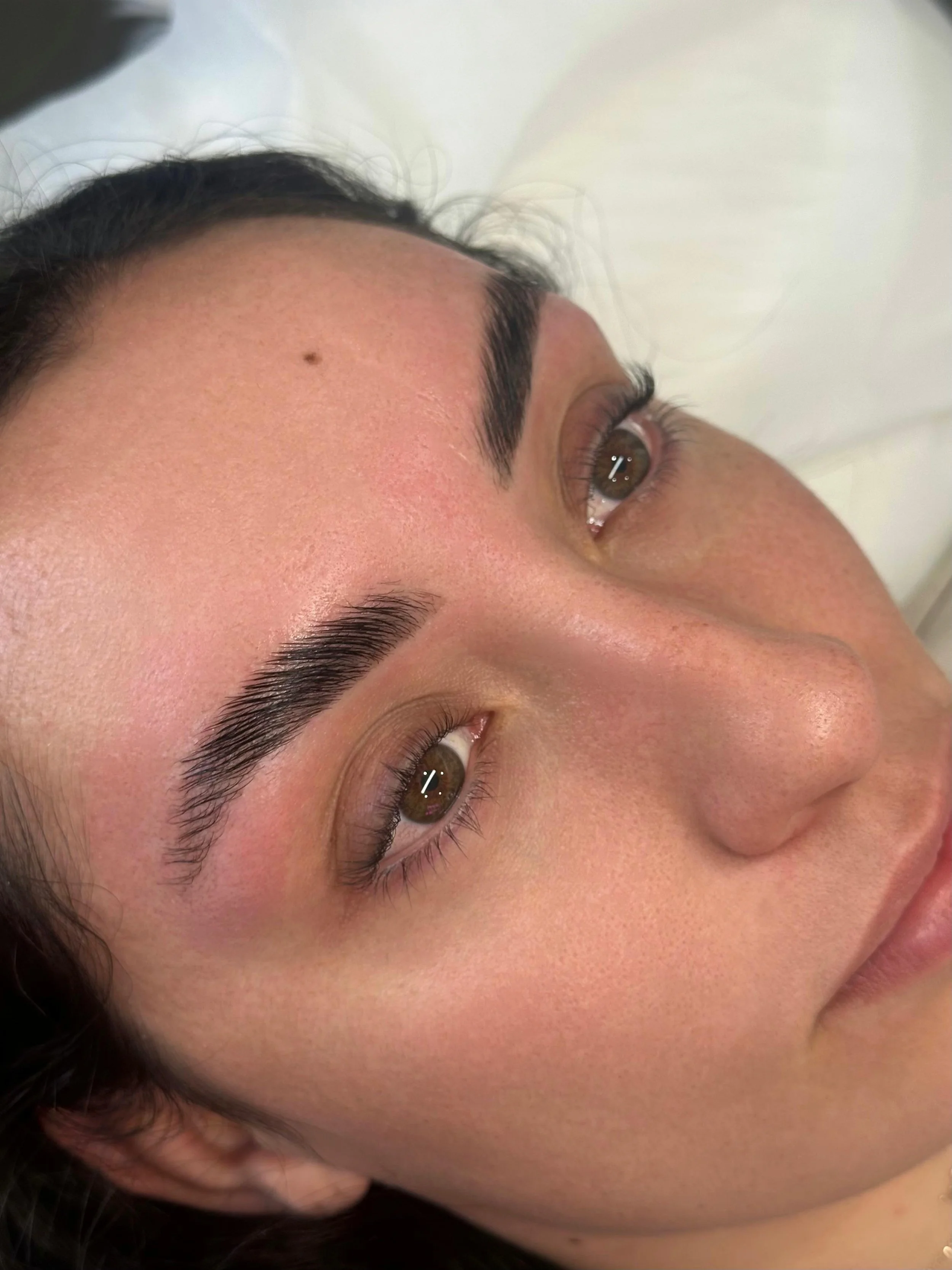 Close-up of a woman's face lying down, focusing on her brown eye, eyebrow, and forehead, with smooth skin and dark hair.