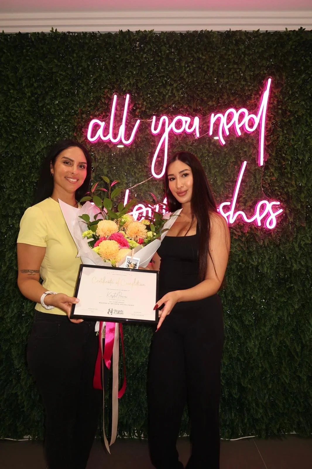 Two women standing in front of a green wall with neon pink sign that says 'all you need is lashes'. The woman on the left has dark hair, is smiling, and is holding a framed certificate and a bouquet of flowers. The woman on the right has long dark hair, is smiling, and is holding the certificate and flowers.