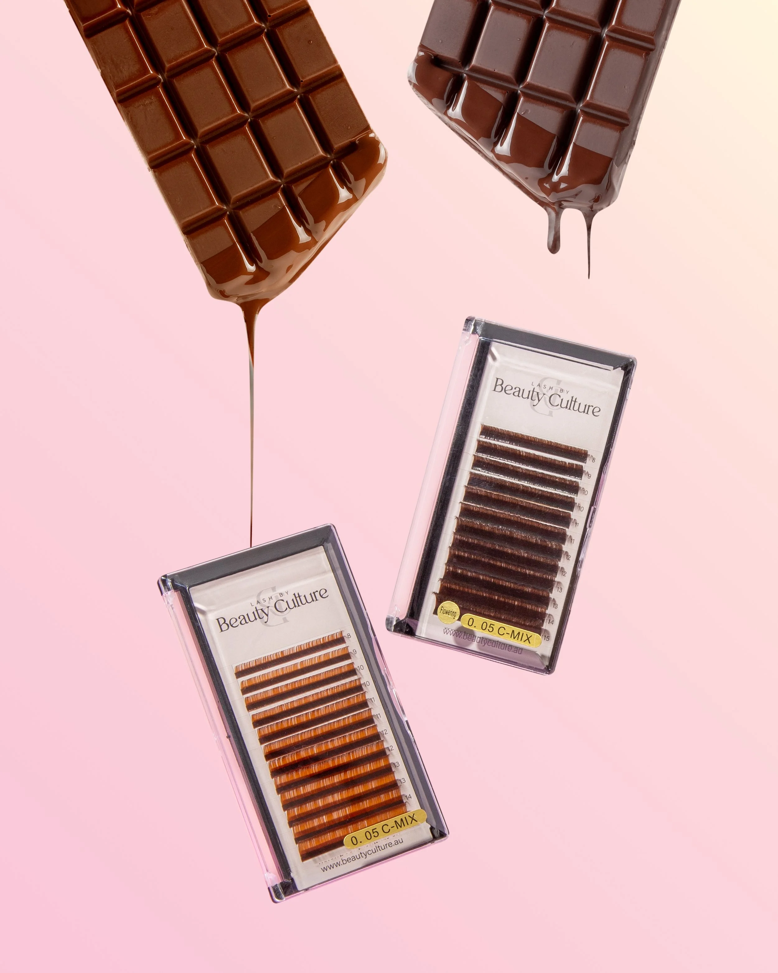 Two broken chocolate bars with melting chocolate drips above two packages of eyelash extensions on a pink background.