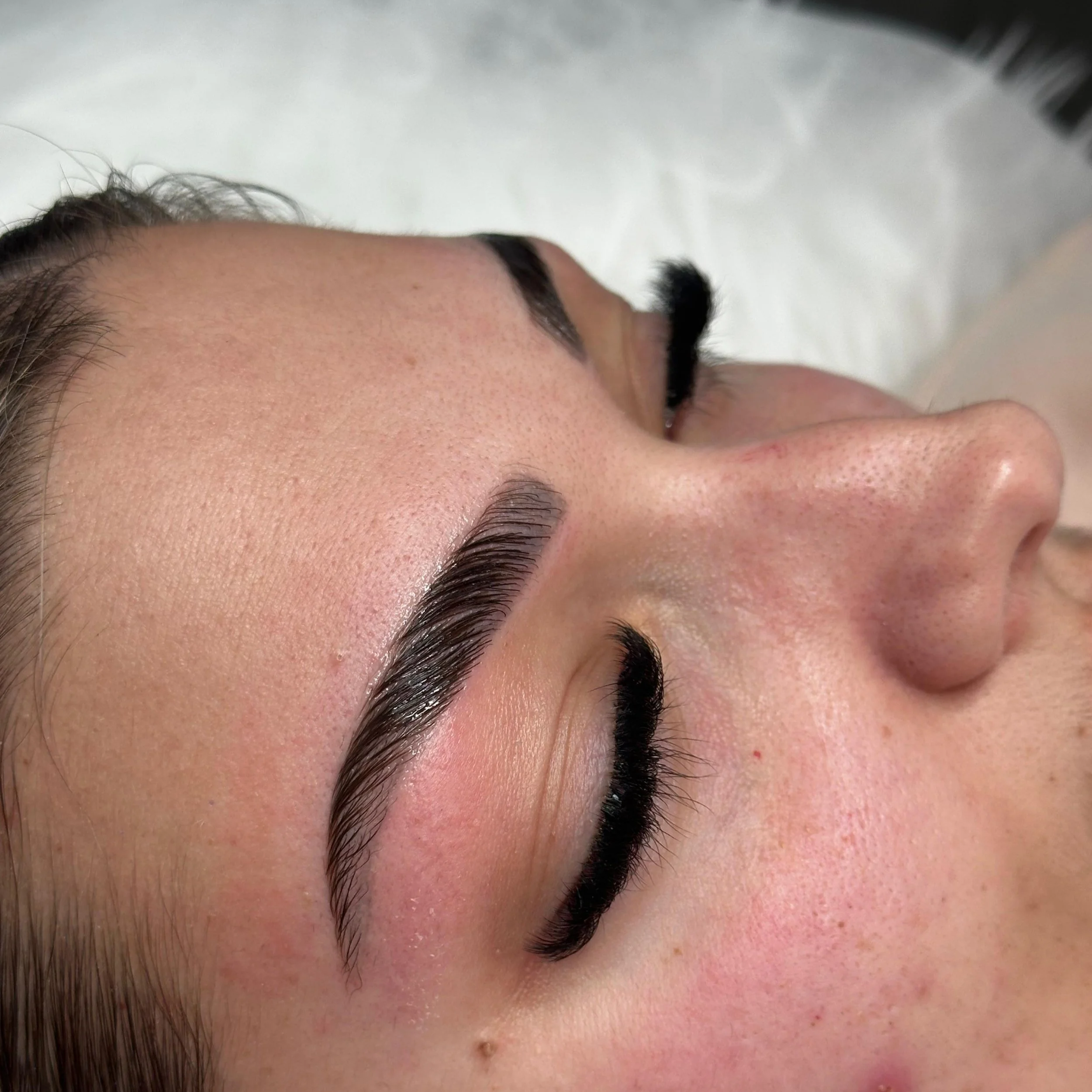 Close-up of a woman's face with freshly applied eyebrow makeup, lying down with eyes closed, showing well-groomed eyebrows, long eyelashes, and natural skin texture.