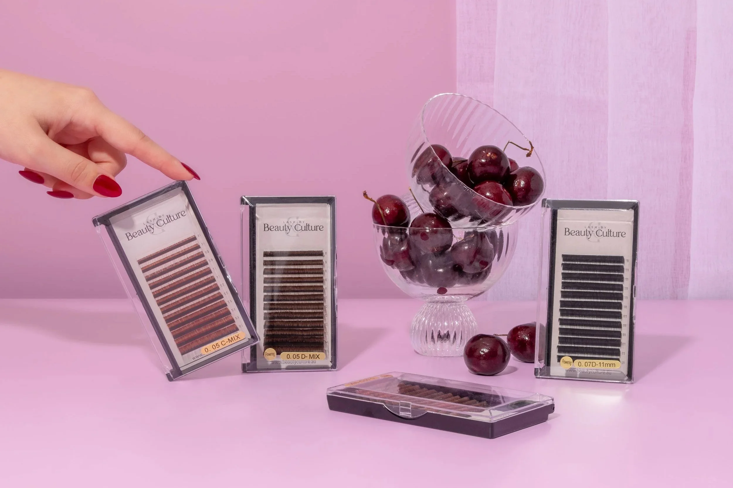 A hand with red painted nails pointing at a box of eyelash extensions on a pink surface. Three more boxes of eyelash extensions and a glass bowl filled with cherries are also on the surface, with some cherries outside the bowl.