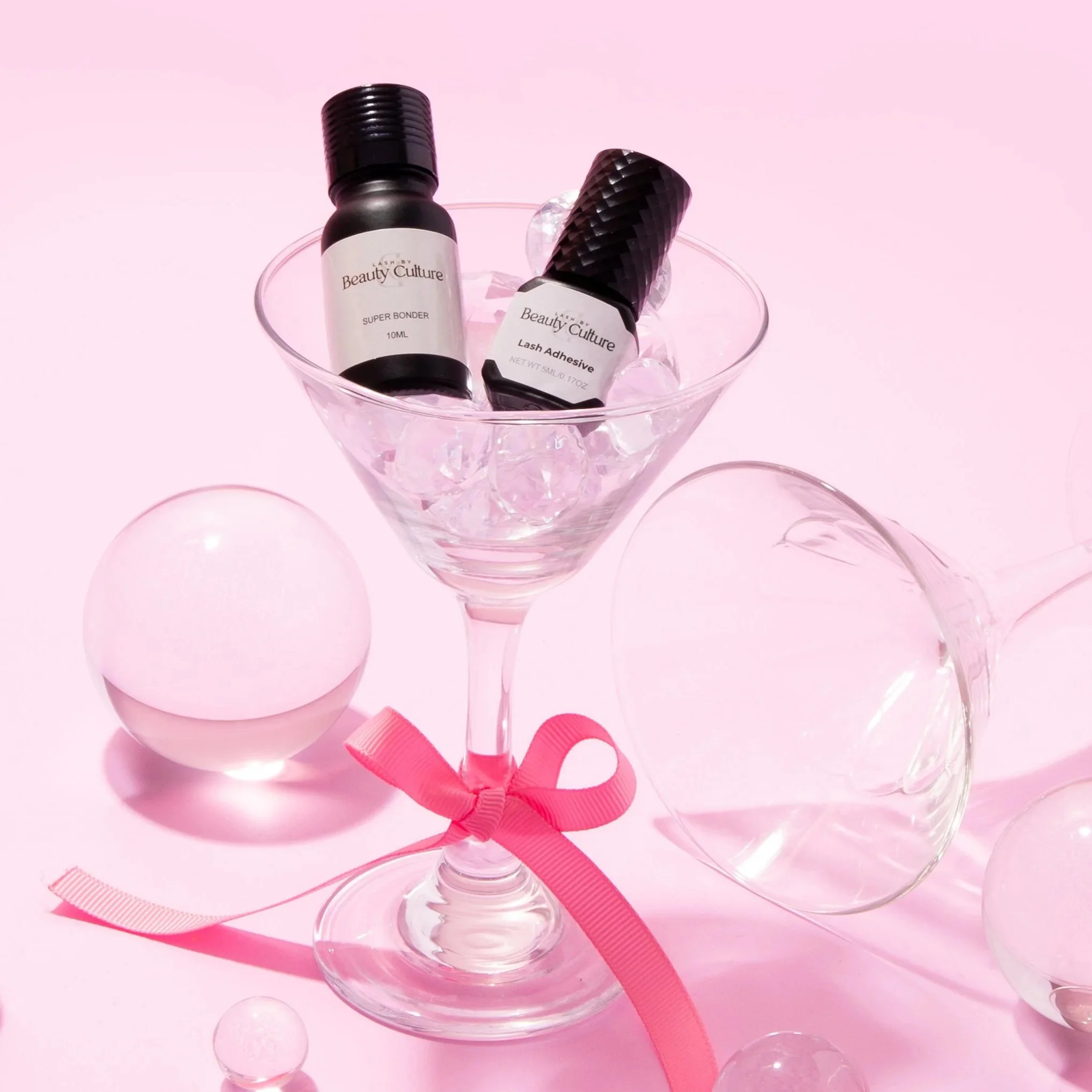 Cosmetic products inside a martini glass filled with ice, with the bottles labeled 'Super Bonder' and 'Lash Adhesive.' The scene is decorated with pink ribbons, glass balls, and a pink background.