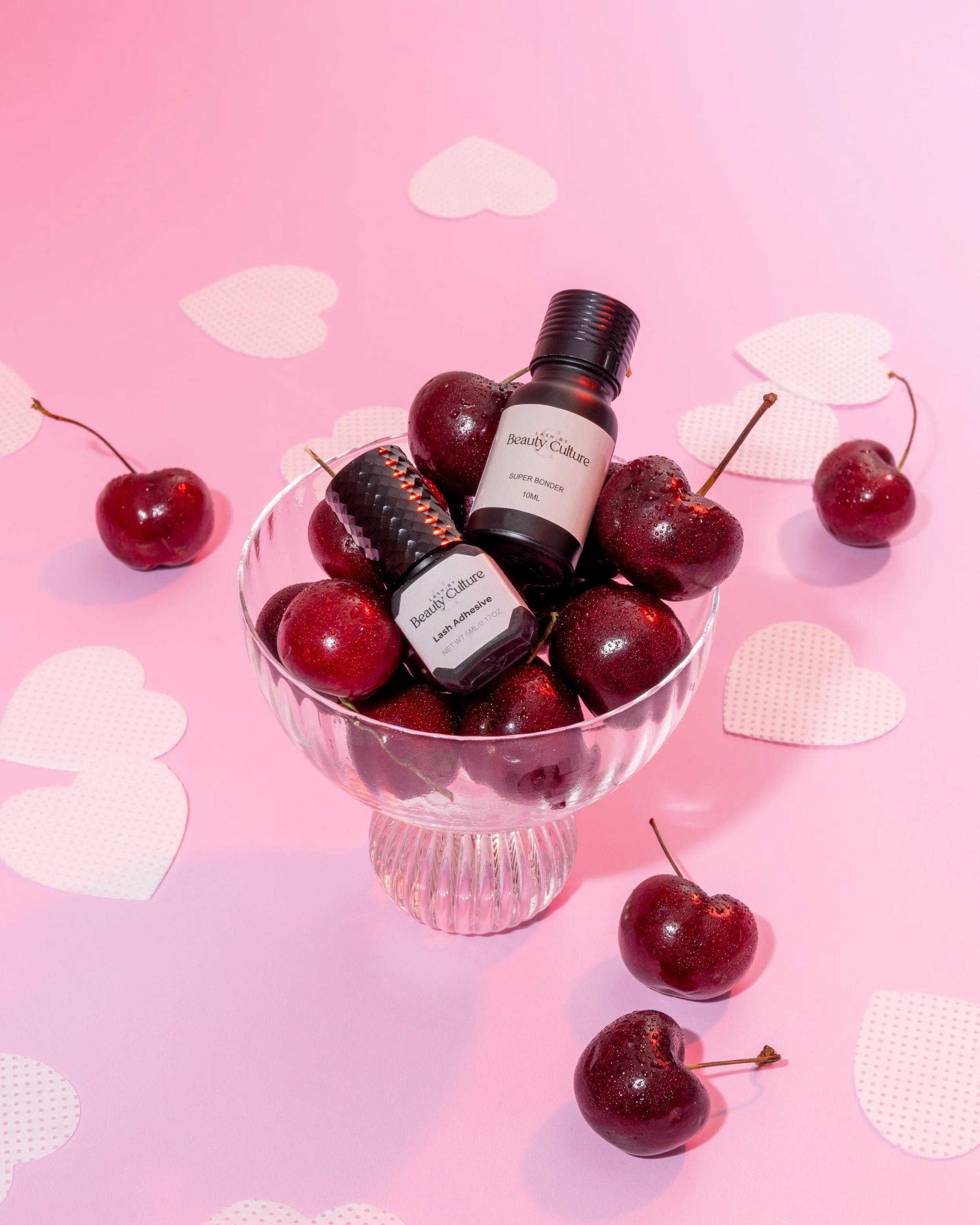 A glass bowl filled with cherries and two bottles of Beauty Culture cosmetic products, set on a pink background with paper heart confetti scattered around.