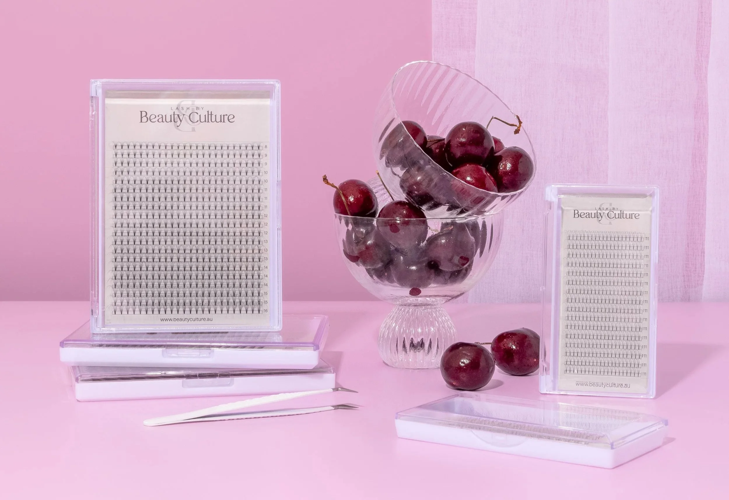 A pink background with a glass bowl filled with cherries, two candied cherries, and several tied cherry stems, surrounded by two rectangular boxes of eyelash extensions, one open and one closed, with tweezers in front.
