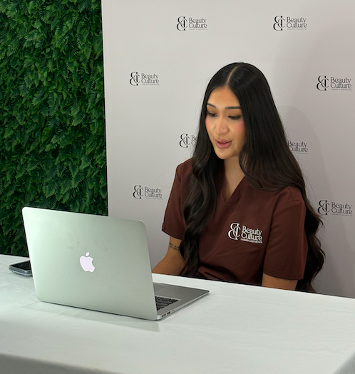 Woman with long dark hair sitting at a white table with a silver MacBook laptop, wearing a brown uniform shirt with a 'Beauty & Culture' logo, against a backdrop with similar logos and a green plant wall.